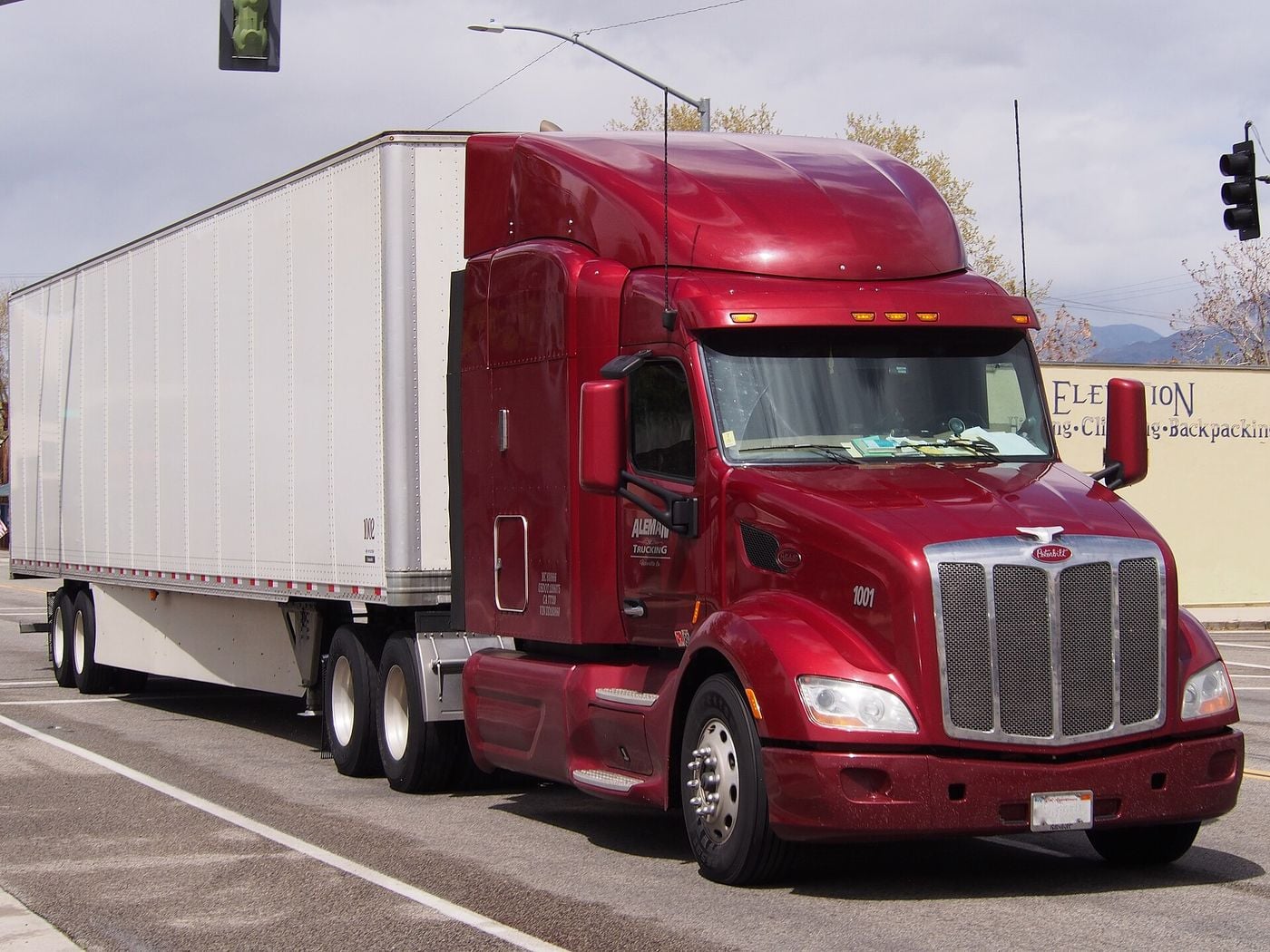 Dark red Peterbilt 579 sleeper tractor with a roof fairing, chrome grille and two rear axles, pulling a white dry-van semi-trailer along the main street of Lone Pine, California, photographed in April 2018