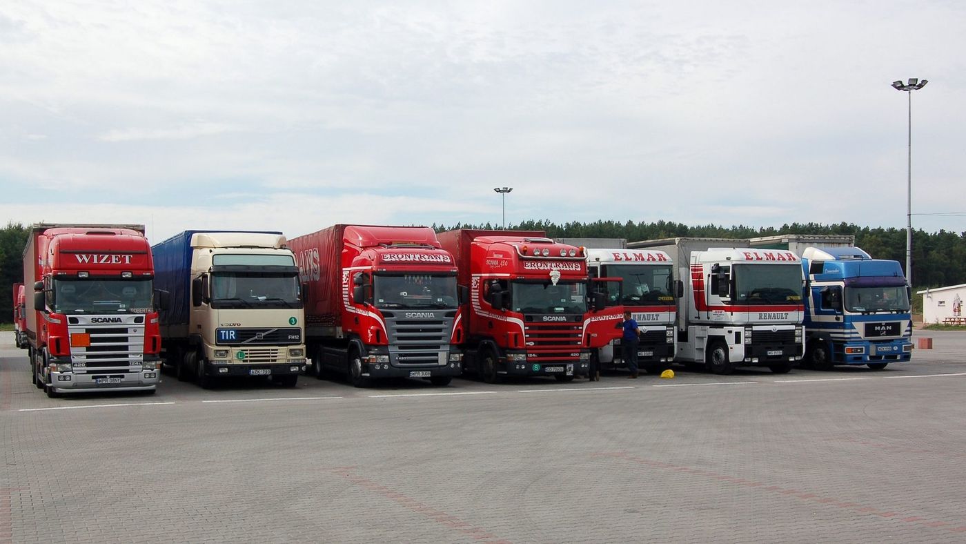 Siedem ciągników siodłowych z naczepami zaparkowanych w rzędzie na placu postojowym kompleksu Nevada Center w Poźrzadle (woj. lubuskie) – m.in. Scania, Volvo z tablicą TIR, Renault i MAN; przy jednej z kabin stoi mężczyzna, w tle las i maszty oświetleniowe, niebo zachmurzone