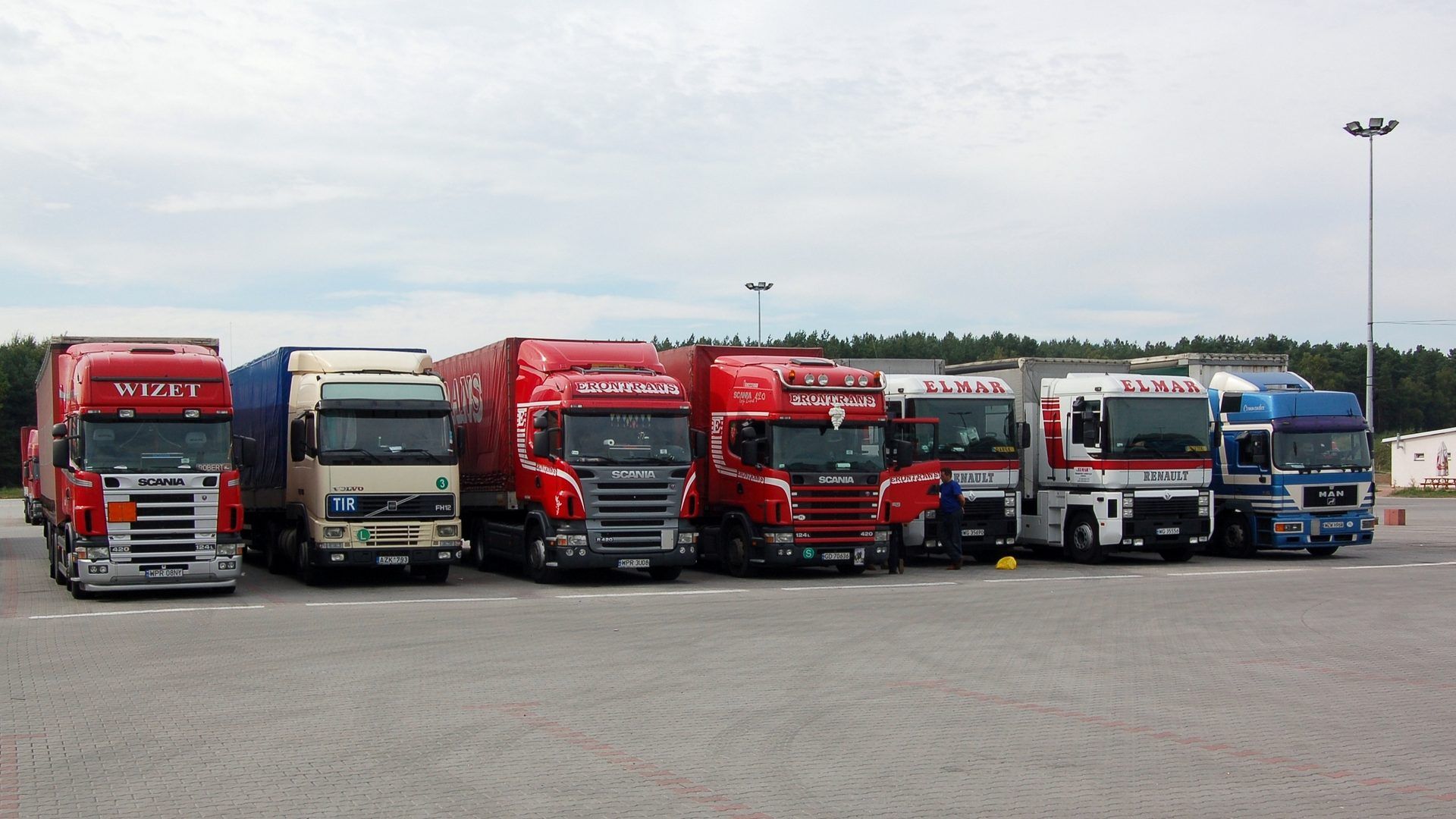 Siedem ciągników siodłowych z naczepami zaparkowanych w rzędzie na placu postojowym kompleksu Nevada Center w Poźrzadle (woj. lubuskie) – m.in. Scania, Volvo z tablicą TIR, Renault i MAN; przy jednej z kabin stoi mężczyzna, w tle las i maszty oświetleniowe, niebo zachmurzone