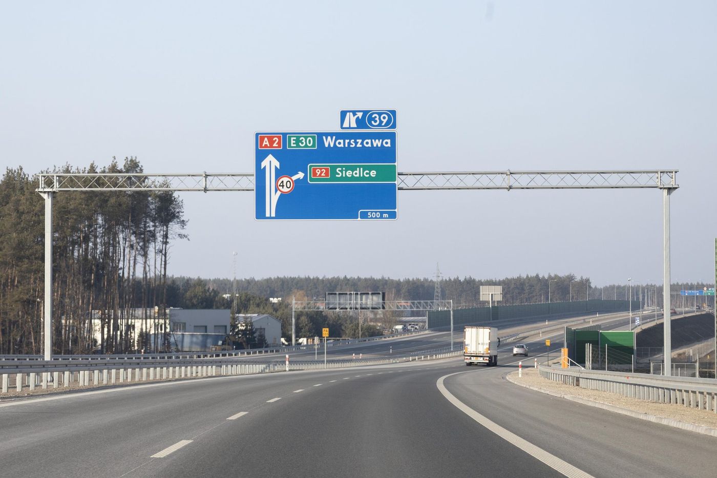 Autostrada A2 koło Siedlec, odcinek Siedlce Zachód – Siedlce Południe: nad jezdnią bramownica z drogowskazem „A2 E30 Warszawa” i zjazdem nr 39 na drogę krajową nr 92 do Siedlec za 500 m; przed nami jadą ciężarówka z białą naczepą z tablicą TIR i samochód osobowy, w tle las i ekrany akustyczne