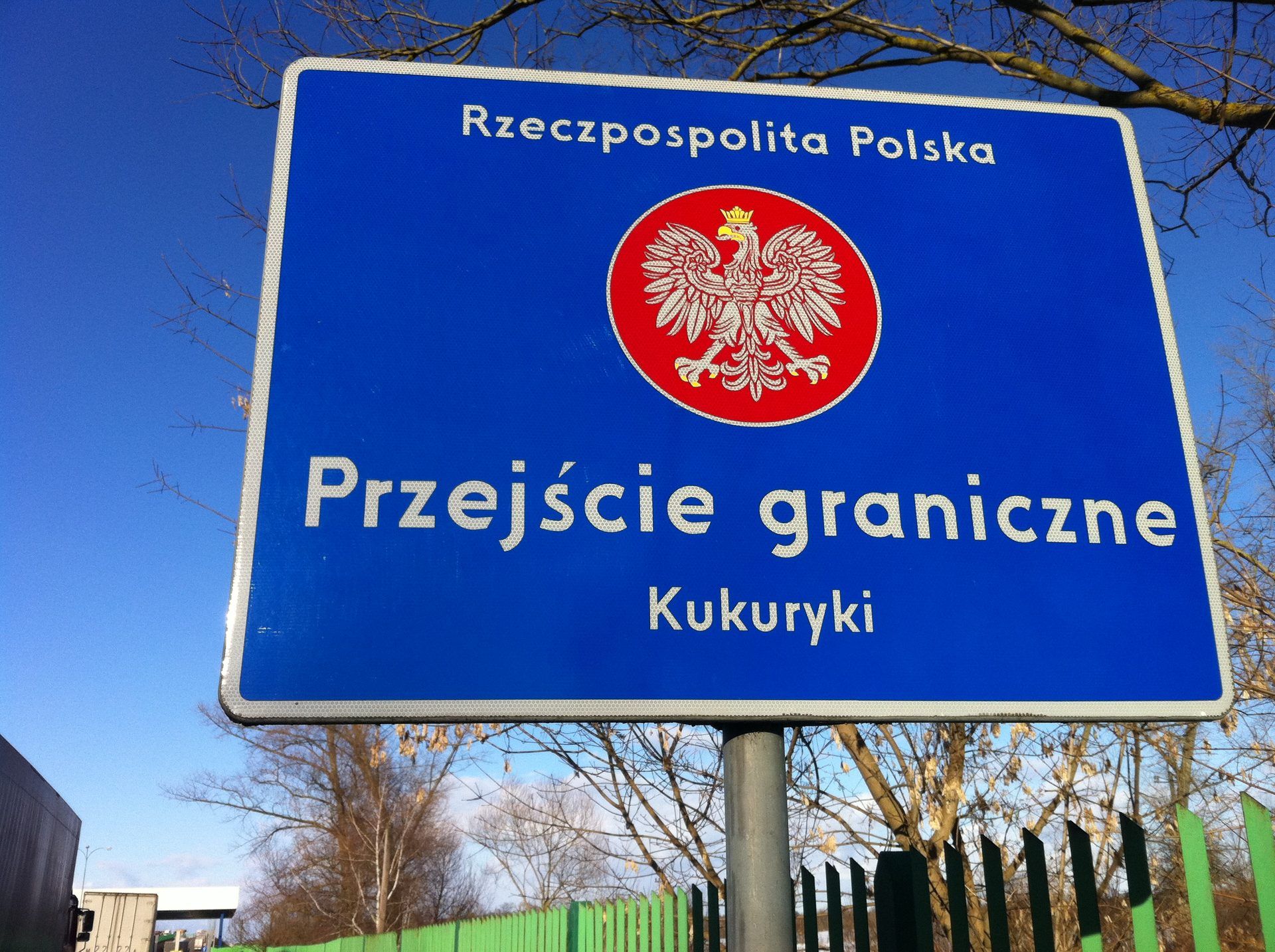 Niebieska tablica z godłem Polski i napisem „Rzeczpospolita Polska. Przejście graniczne Kukuryki” na tle nieba; przy lewej krawędzi kadru fragmenty ciężarówek, w tle zadaszenie przejścia granicznego i zielone ogrodzenie