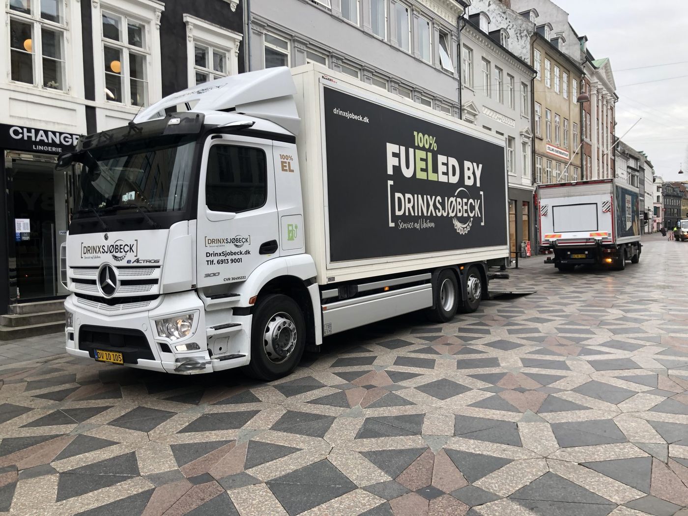 Biała elektryczna ciężarówka Mercedes-Benz eActros 300 z zabudową kontenerową i napisem „100% EL” na kabinie stoi na wybrukowanym deptaku w Kopenhadze; dalej druga ciężarówka dostawcza