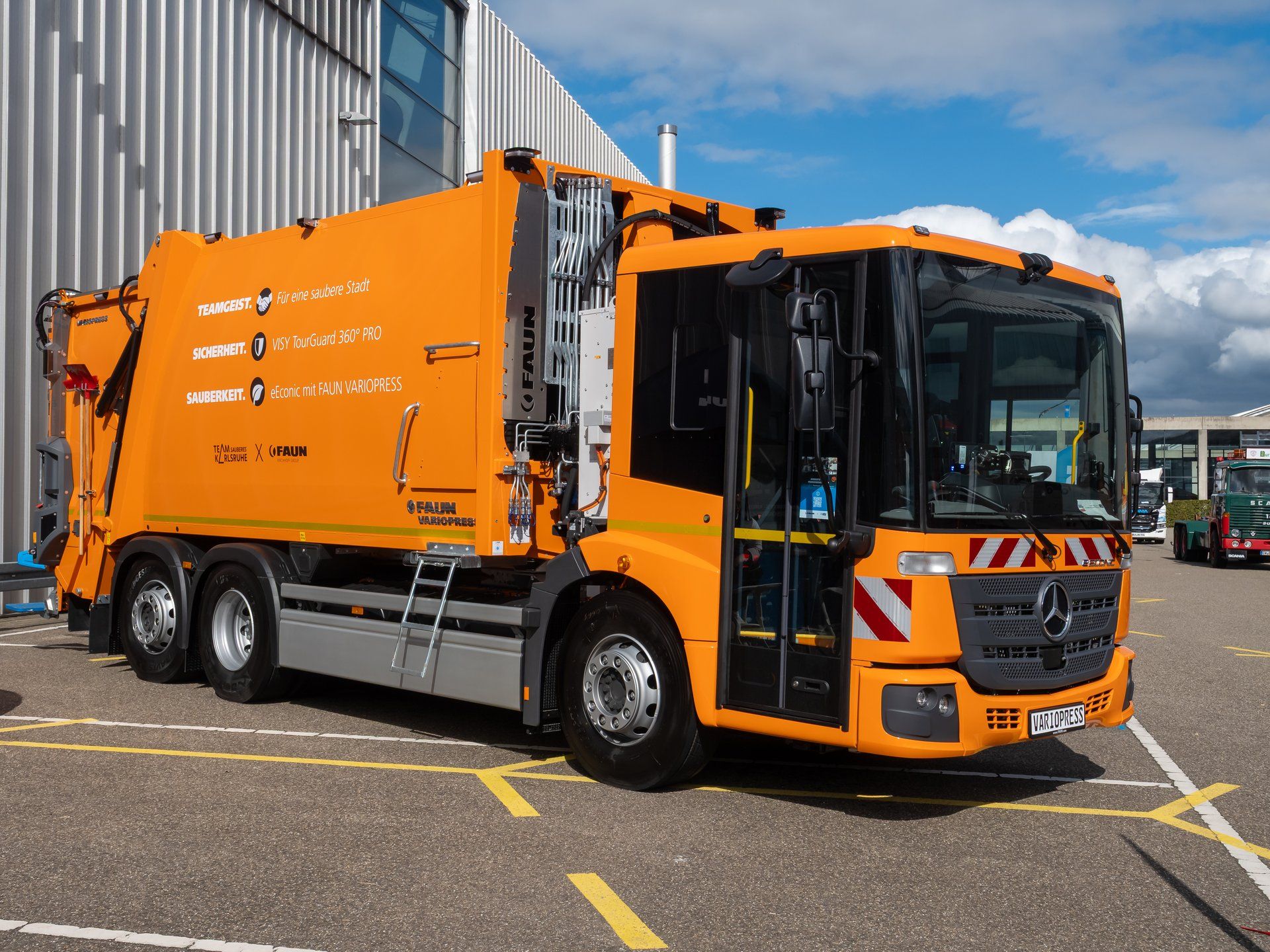 Orange Mercedes-Benz eEconic electric refuse collection vehicle with a FAUN VARIOPRESS rear-loader body and a glazed folding cab door, at the Nufam 2025 trade fair in Rheinstetten