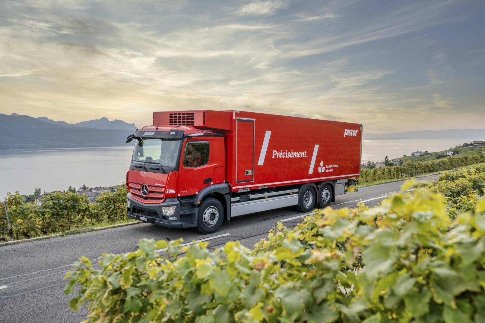 Red electric Mercedes-Benz box truck driving along a narrow road beside terraced vineyards, with a lake and mountains behind it