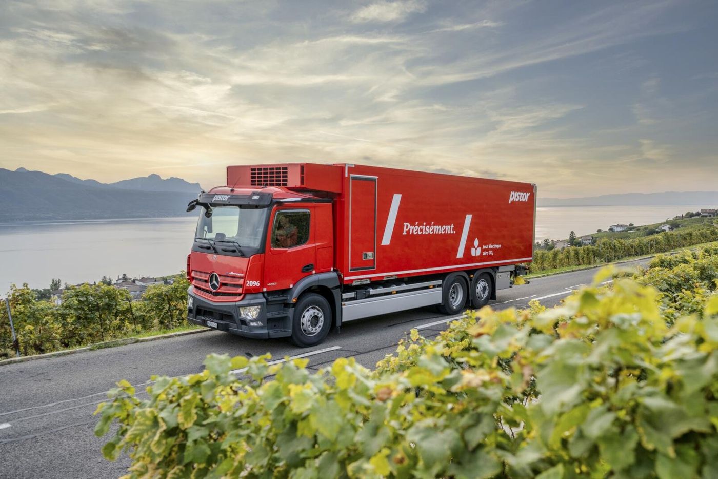 Red electric Mercedes-Benz box truck driving along a narrow road beside terraced vineyards, with a lake and mountains behind it