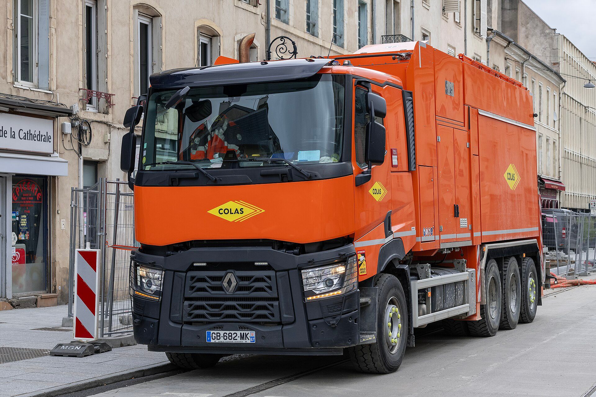 Orange Renault Trucks C 520 with a single front axle, three rear axles and a suction-excavator body in Colas livery, parked at roadworks on a town street