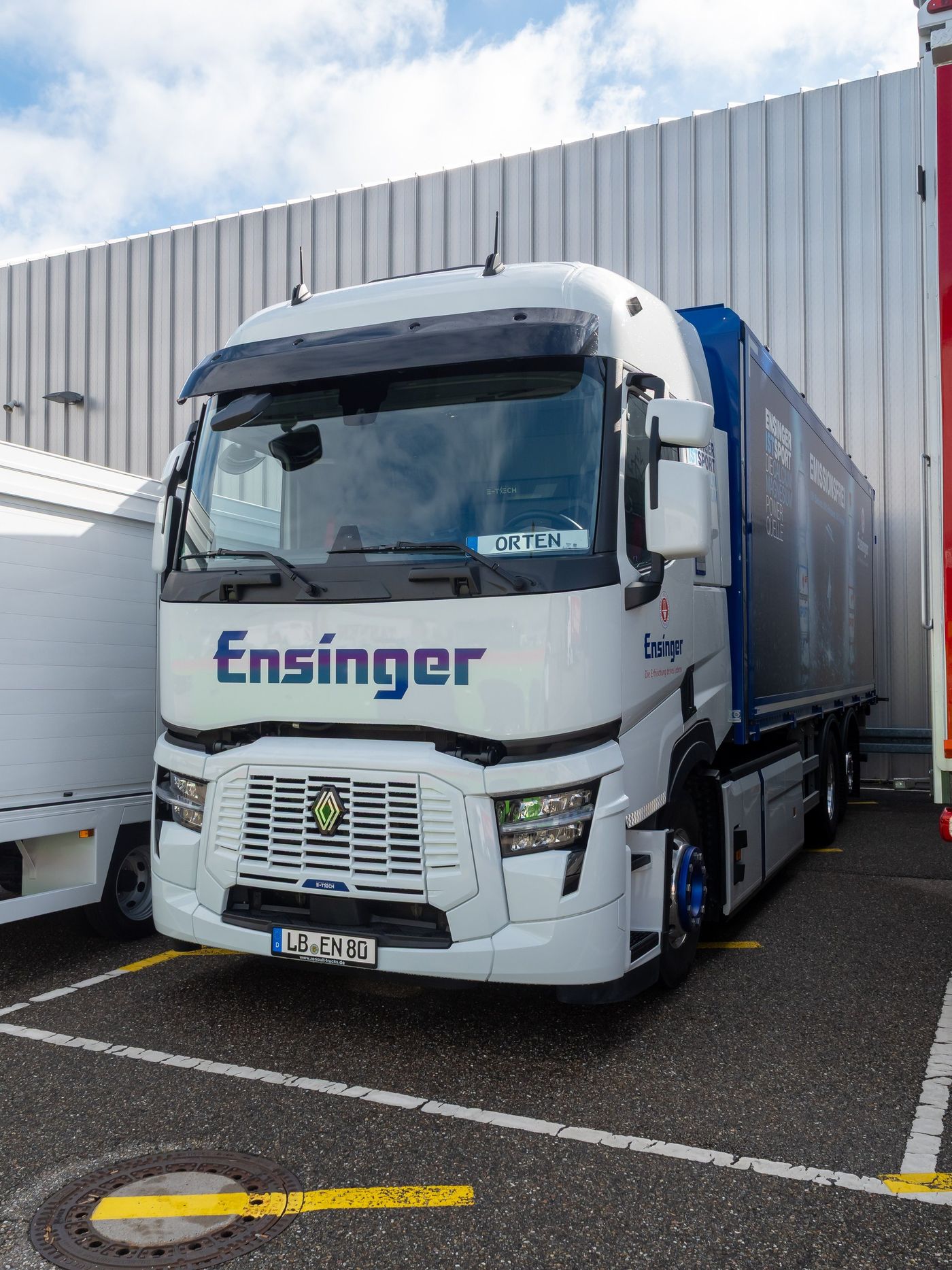 White three-axle Renault Trucks E-Tech T rigid in Ensinger livery, with a closed louvred grille, an E-Tech badge beneath it and a blue-framed load body lettered Emissionsfrei, parked beside a metal-clad hall