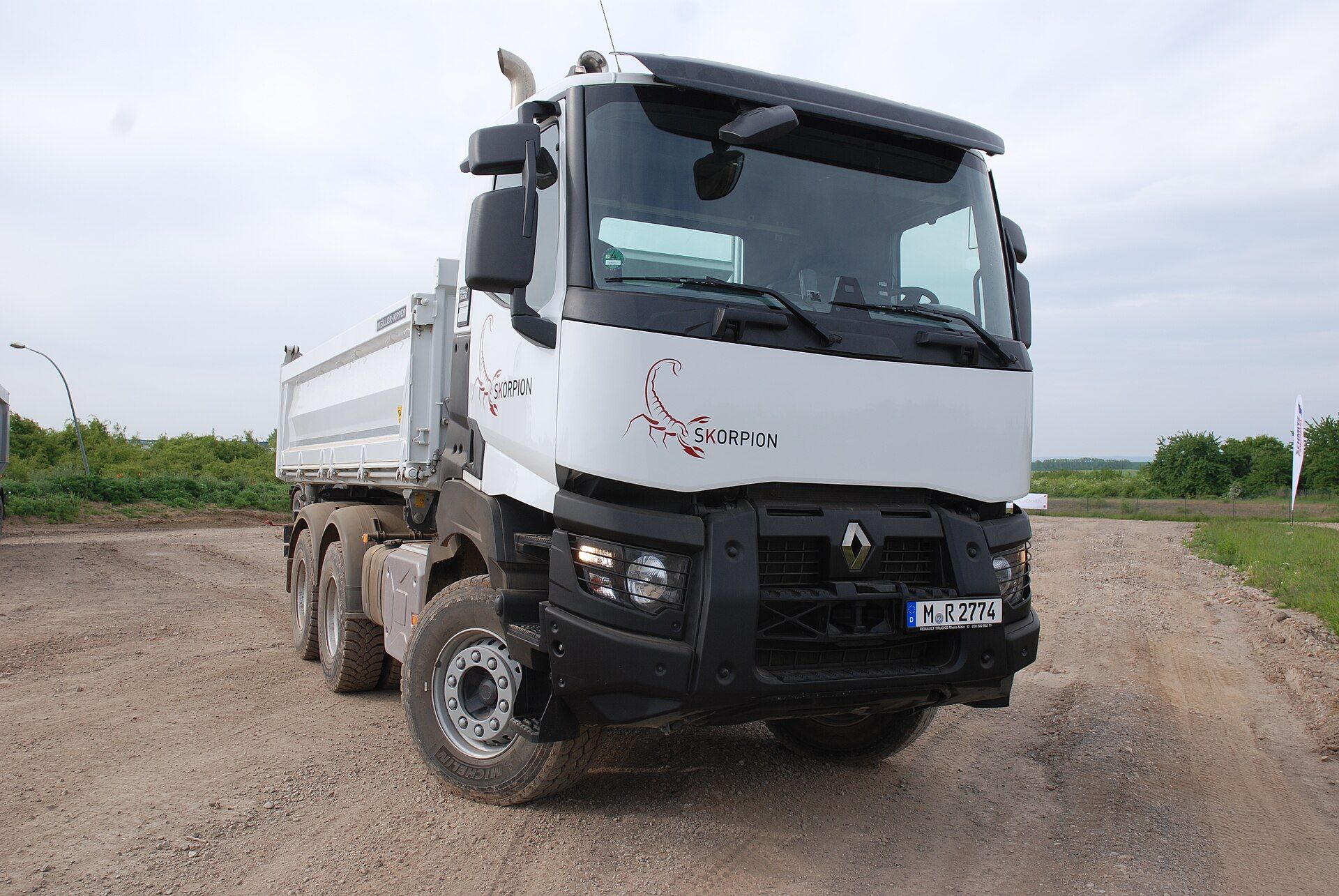 White three-axle Renault Trucks K tipper with the earlier front, larger headlamp units behind grilles and a black steel bumper, standing on a gravel test ground with a scorpion decal on the cab