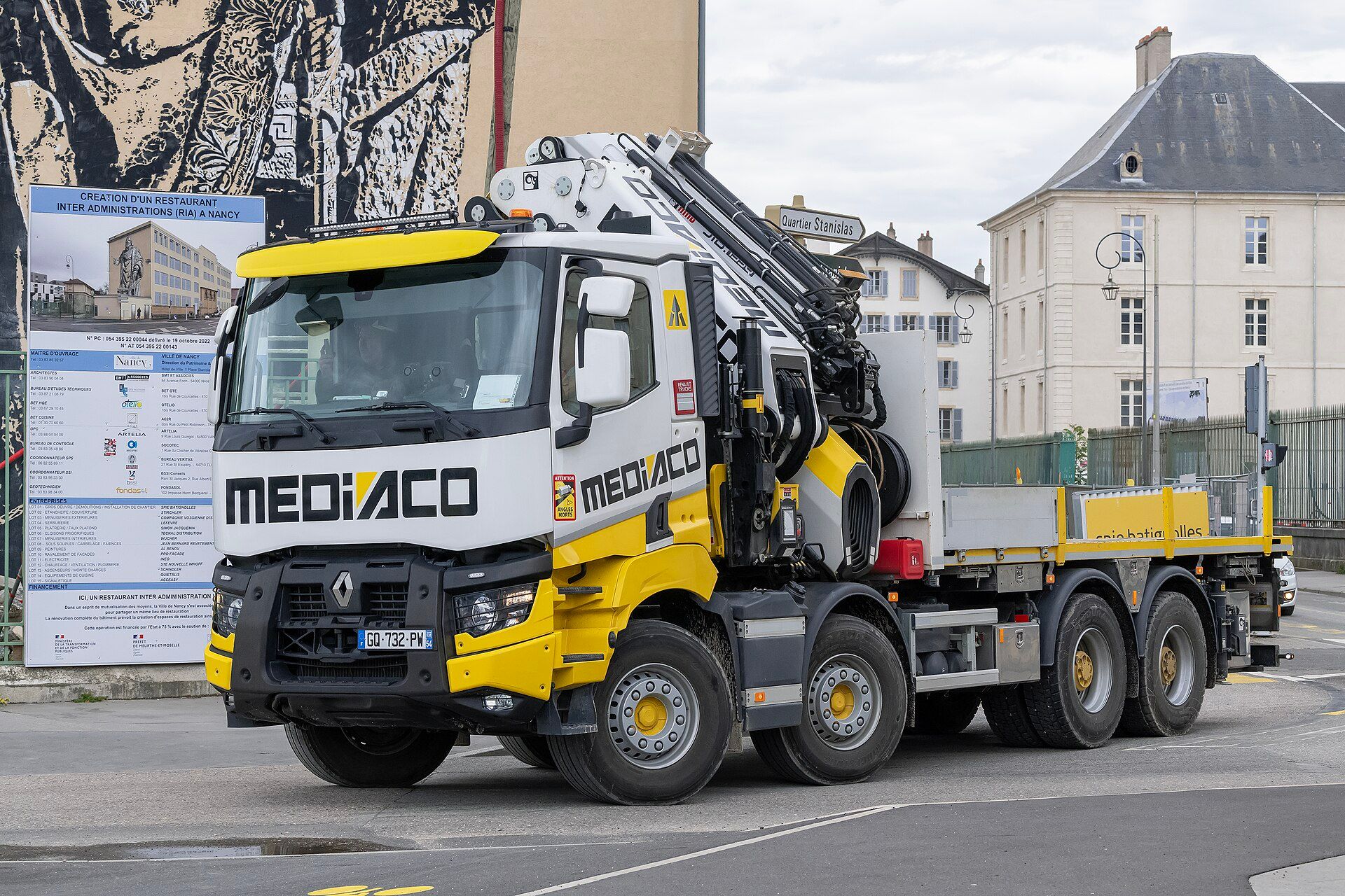 White and yellow Renault Trucks K 480 with four axles, two at the front and two at the rear, carrying a loader crane behind the cab and a flatbed body, on a town street beside a building-site signboard