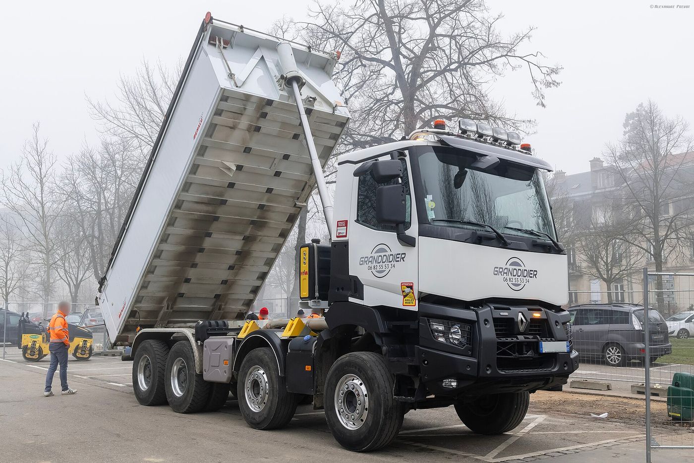 White Renault Trucks K 460 four-axle tipper, two axles at the front and two at the rear, with a black steel bumper and grilles over the headlamps, its body raised at a roadworks site on a foggy day with bare trees behind