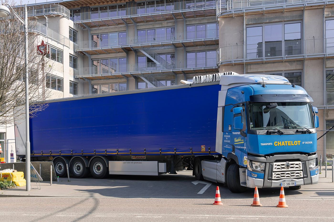 Niebieski dwuosiowy ciągnik siodłowy Renault Trucks T w wersji sprzed 2021 r. – chromowane listwy atrapy chłodnicy, pełny chromowany romb i oddzielne reflektory – z naczepą kurtynową, przy krawężniku w mieście