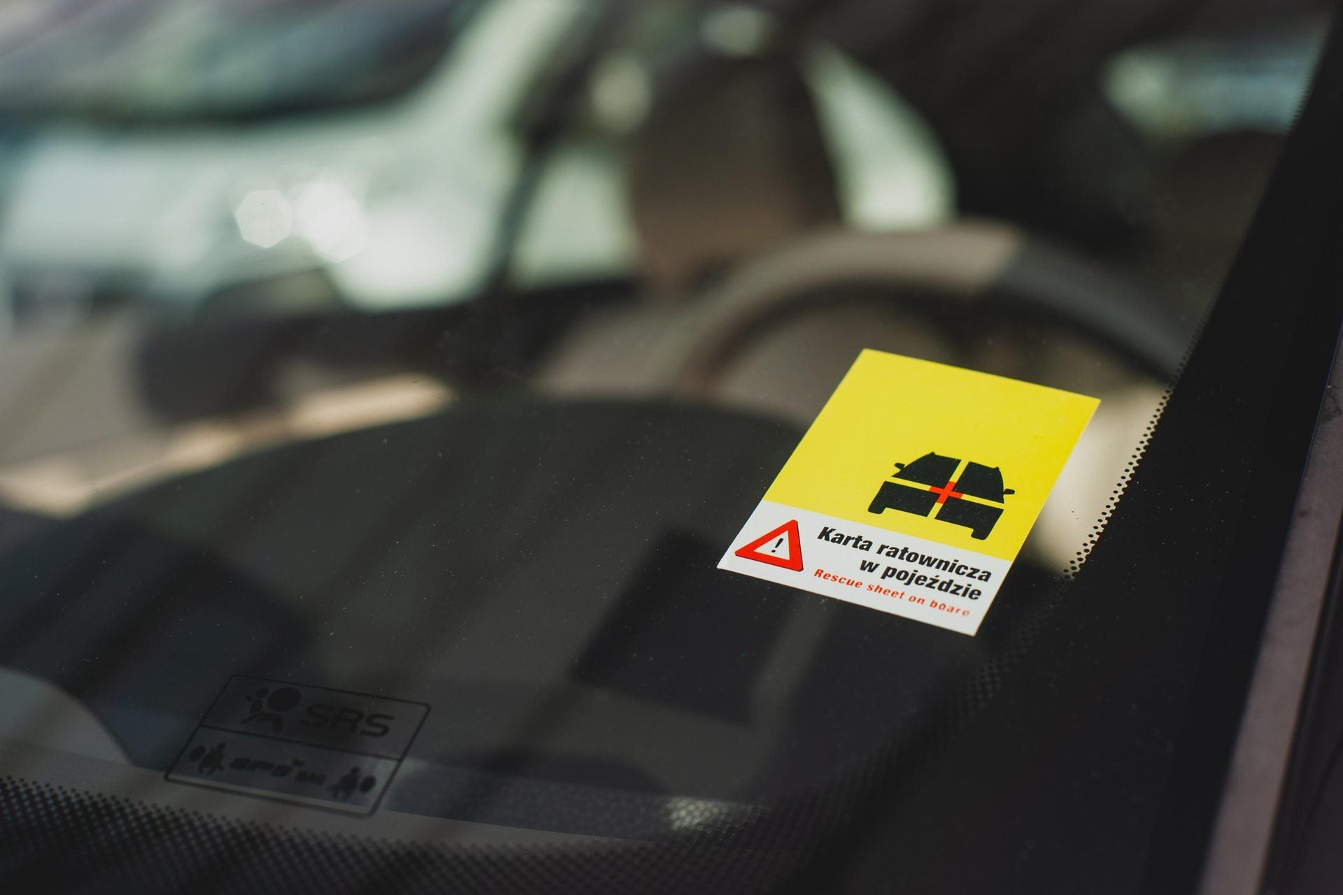 Yellow and white sticker on the inside of a car windscreen reading Karta ratownicza w pojeździe and Rescue sheet on board, with a black vehicle pictogram and a red warning triangle