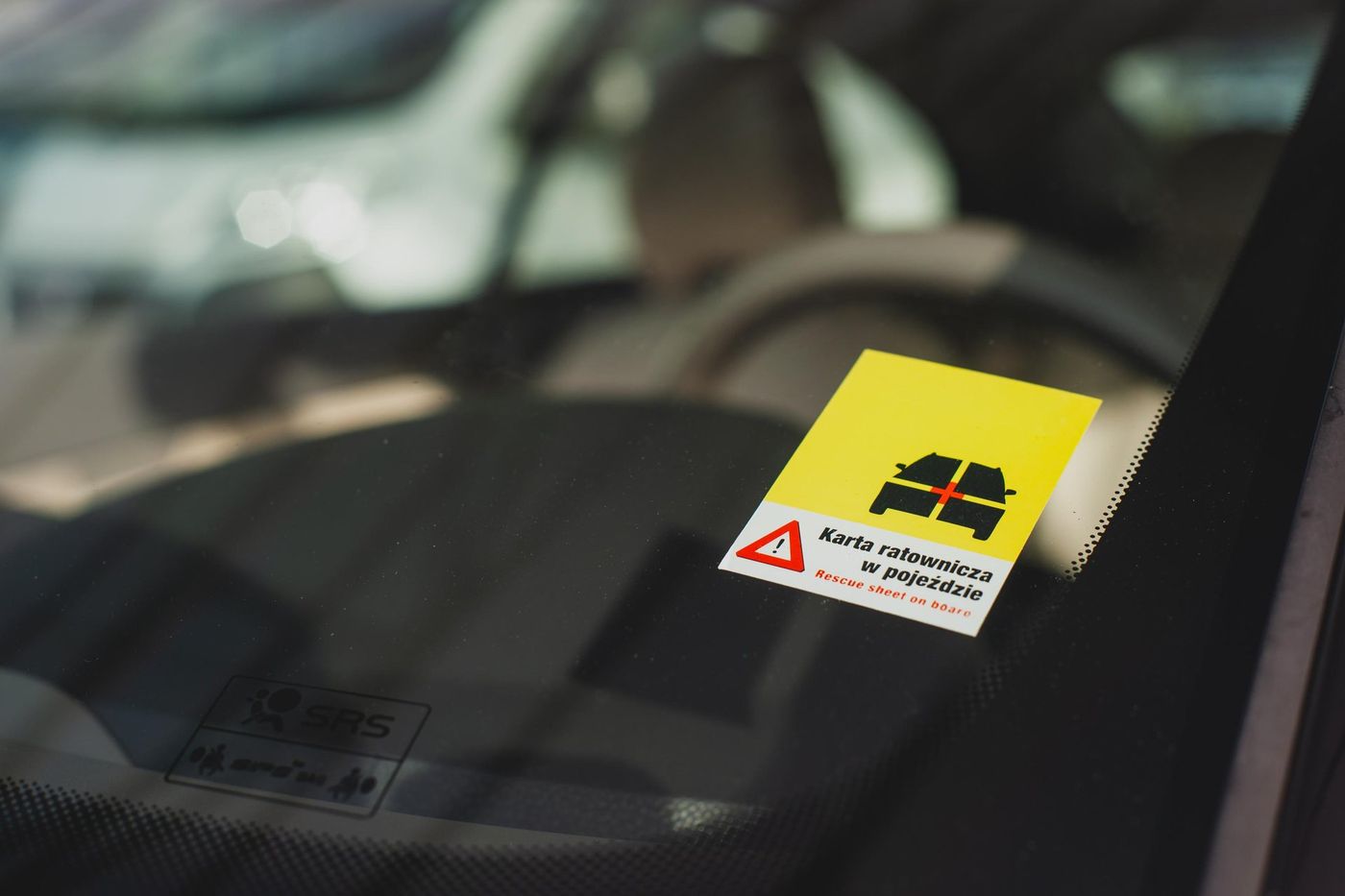 Yellow and white sticker on the inside of a car windscreen reading Karta ratownicza w pojeździe and Rescue sheet on board, with a black vehicle pictogram and a red warning triangle
