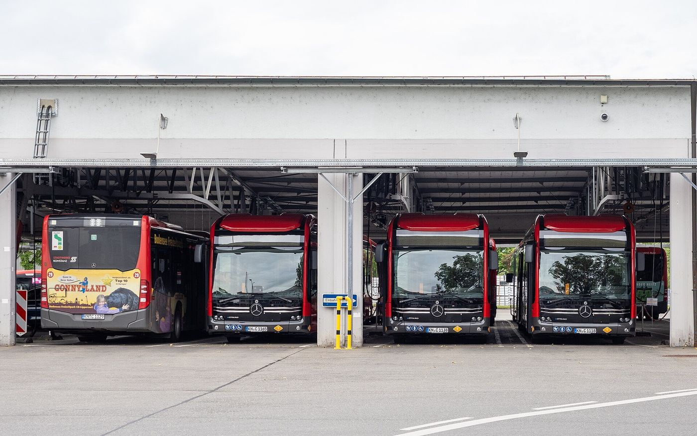 Three red Mercedes-Benz eCitaro city buses parked nose-out in the covered bays of a bus depot, with a fourth red bus seen from behind at the left