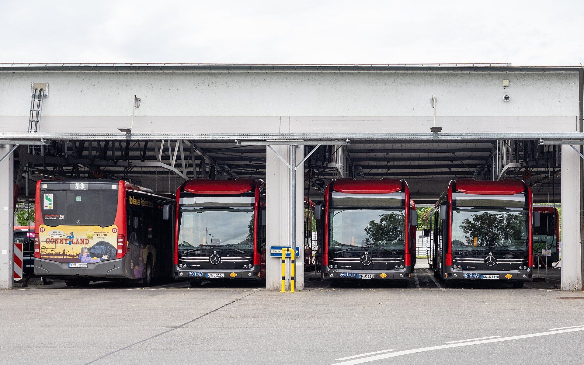 Three red Mercedes-Benz eCitaro city buses parked nose-out in the covered bays of a bus depot, with a fourth red bus seen from behind at the left