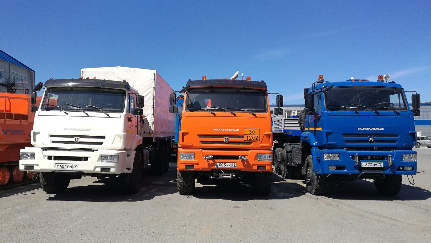 Три грузовика KAMAZ в ряд на площадке в Тюмени: белый с тентованным кузовом, оранжевый с табличкой опасного груза и синий седельный тягач