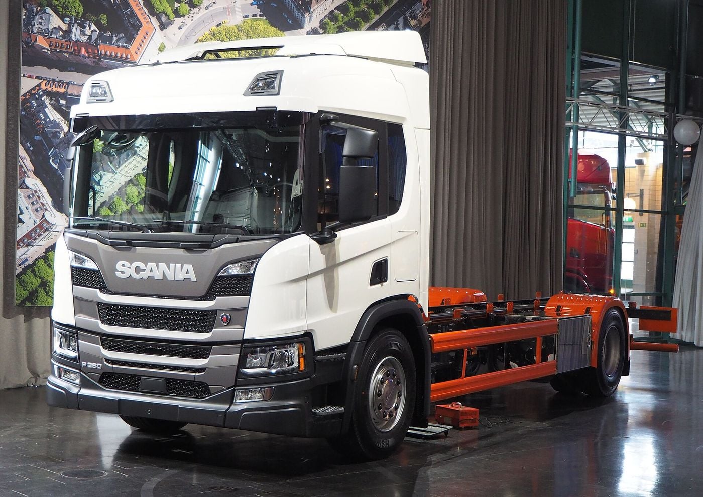 White Scania P 280 two-axle rigid chassis cab with a roof deflector and an orange-painted chassis frame, front three-quarter view on an indoor display floor