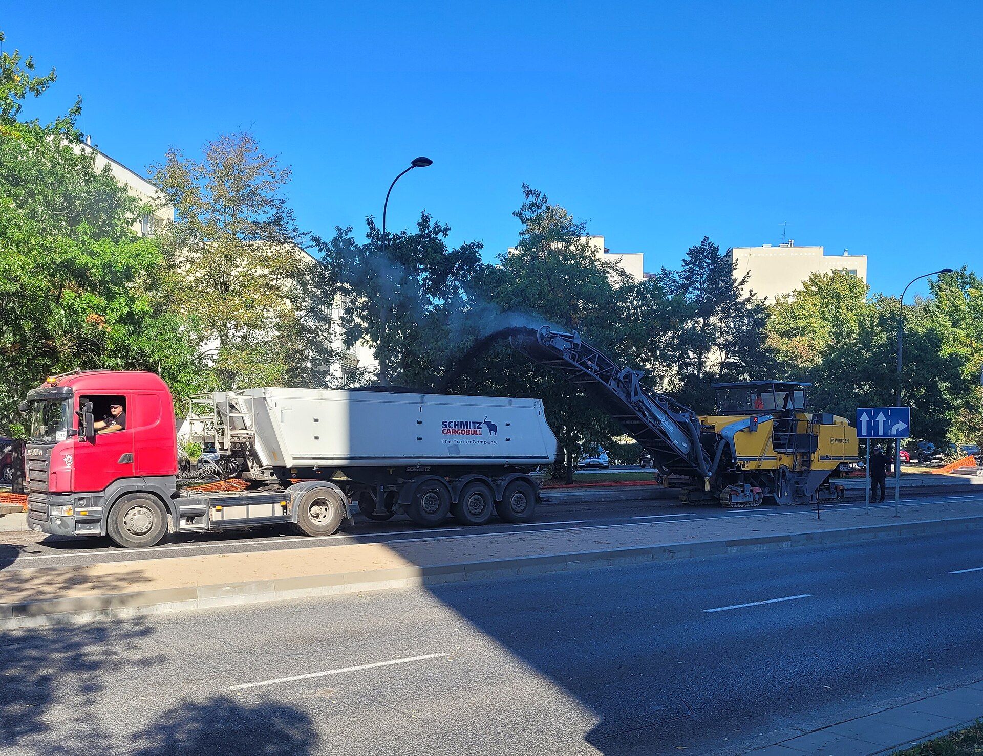 Dreiachsiger Schmitz-Cargobull-Kippsattelauflieger hinter einer roten Scania-Zugmaschine, der bei einer Fahrbahnsanierung in Warschau von einer Kaltfräse mit Fräsgut beladen wird