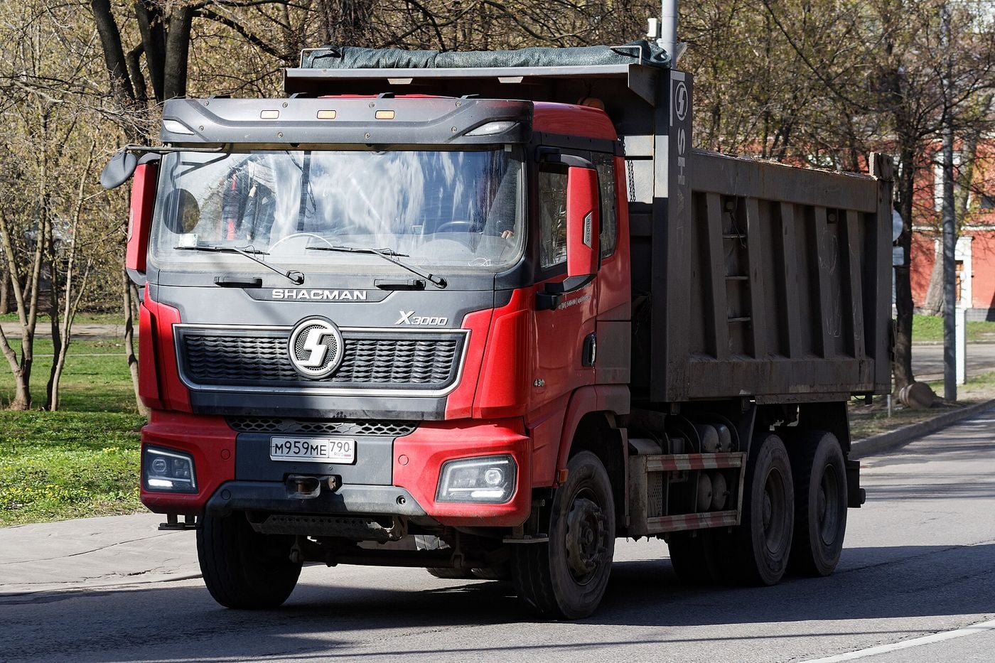 Red SHACMAN X3000 three-axle tipper with a flat-roof cab and a dark grey steel tipper body, driving along a tree-lined street in Moscow in early spring