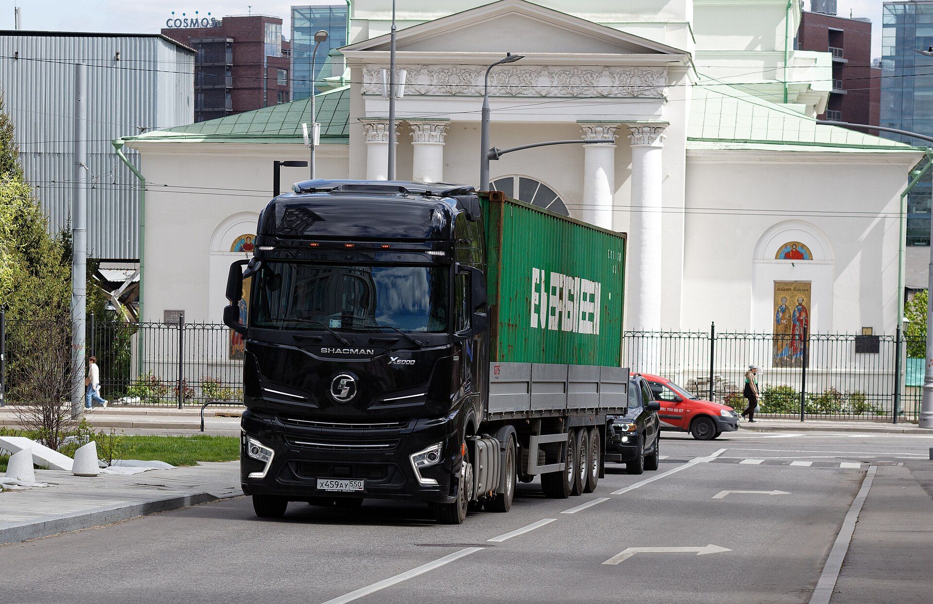 Black SHACMAN X6000 4x2 tractor unit with a high-roof cab, hauling a green shipping container on a three-axle flatbed semi-trailer past a white classical church in Moscow