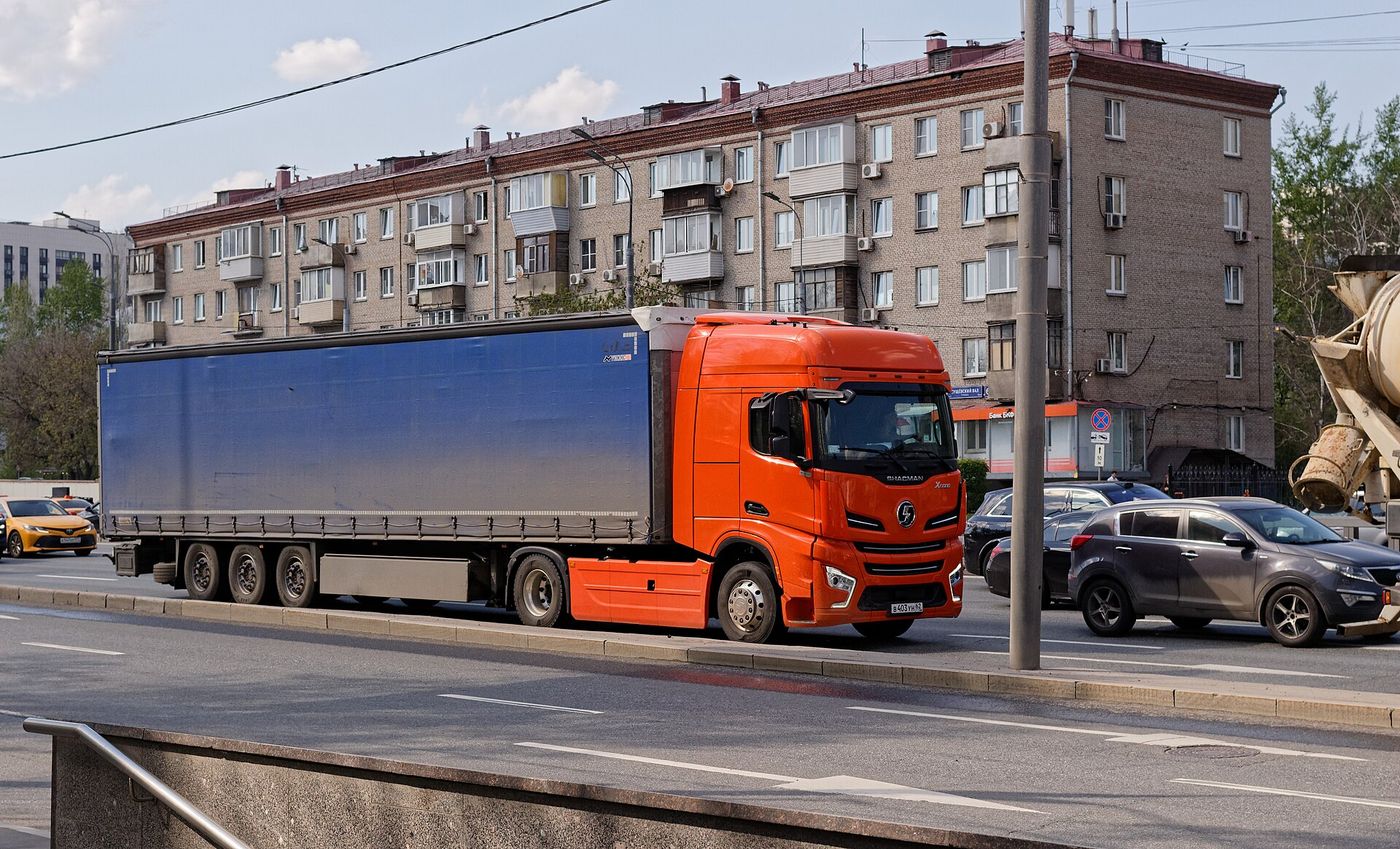 Orange SHACMAN X6000 4x2 tractor unit with a high-roof cab, pulling a blue three-axle curtainsider semi-trailer along a city street in Moscow, with a five-storey brick apartment block behind