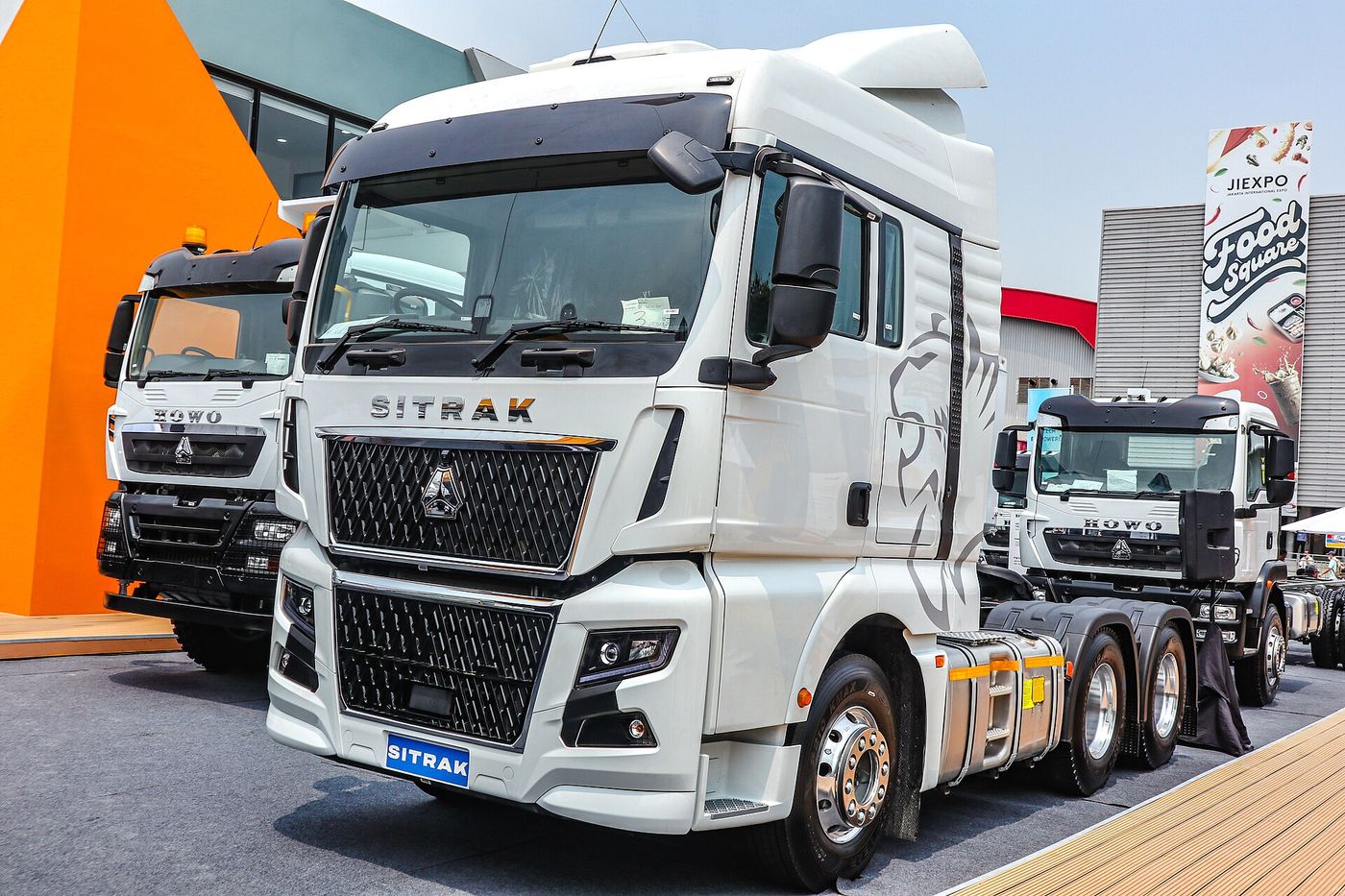 White SITRAK 6x4 tractor unit with a high-roof cab, a large mesh grille and a lion graphic on the cab side, on a show stand at Mining Indonesia 2026 in Jakarta, with HOWO trucks parked on either side