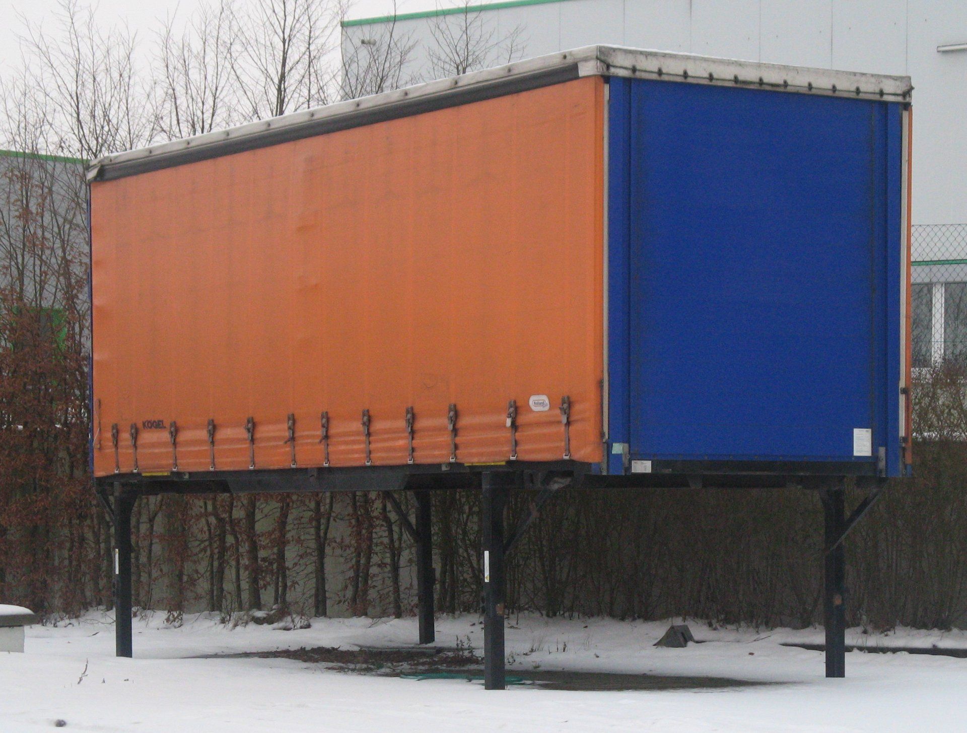 Curtainsider swap body with an orange side curtain and a blue rear curtain, standing on four support legs in a snowy yard