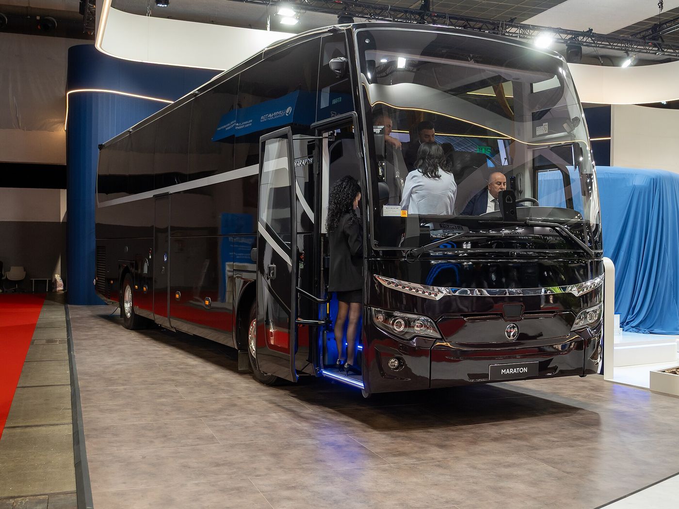 Black Temsa Maraton two-axle coach with a MARATON plate under the grille and its front door open, on the Temsa stand at Busworld Europe 2025 in Brussels