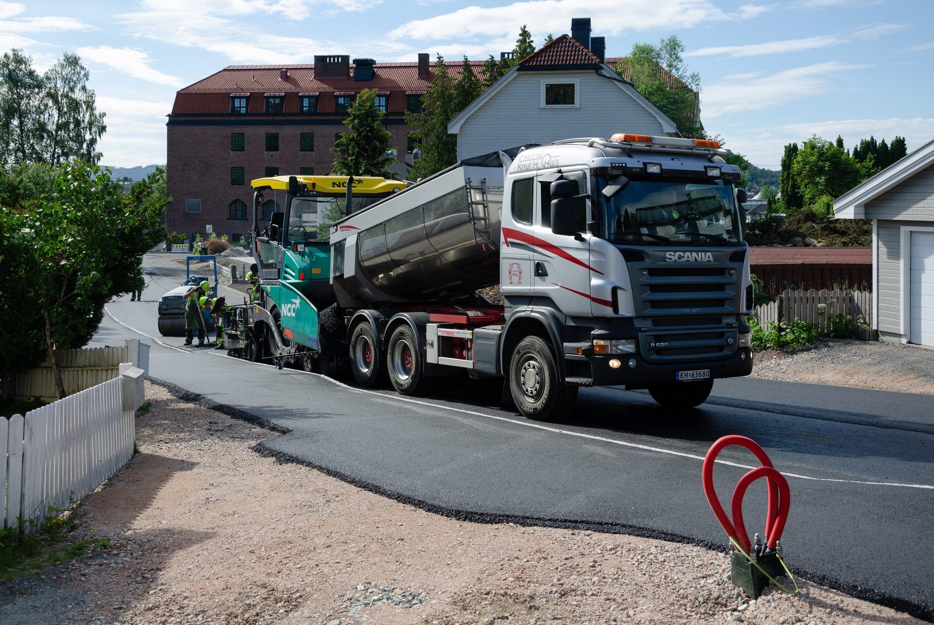 Three-axle Scania tipper with a half-round body raised to feed asphalt into a Vögele road paver on Tomineborgveien in Drammen, Norway, in July 2019