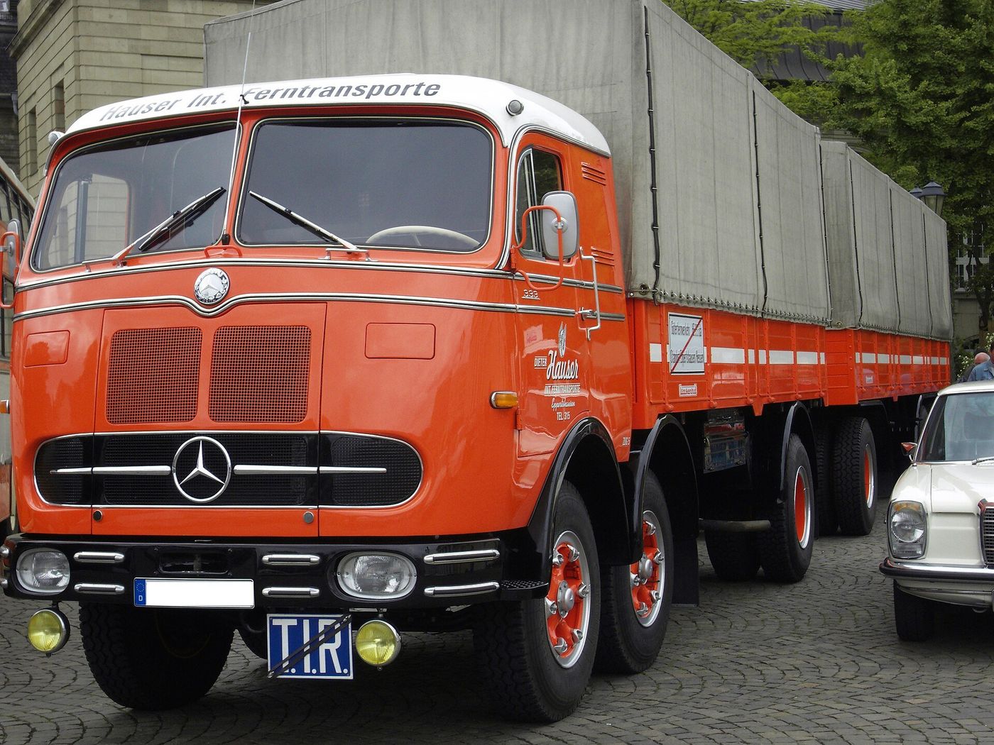 Orange Mercedes-Benz lorry with a full trailer and canvas load cover, with a blue T.I.R. plate on the front