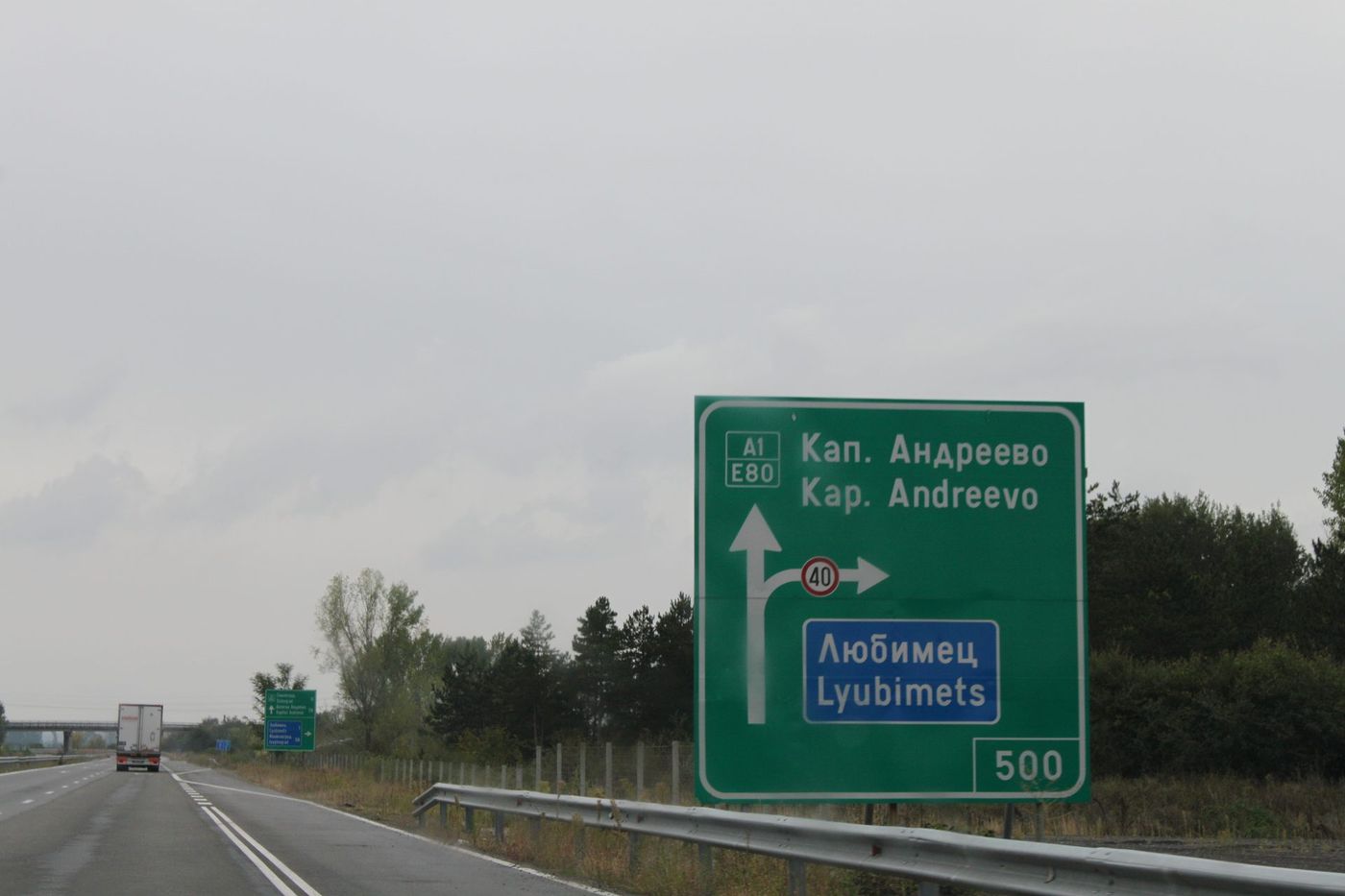 Bulgaristan'da bir otoyolda A1/E80 numaralı, Kapitan Andreevo yönünü ve Lyubimets çıkışını gösteren yeşil tabela; önde kapalı kasalı bir tır ilerliyor, gökyüzü bulutlu
