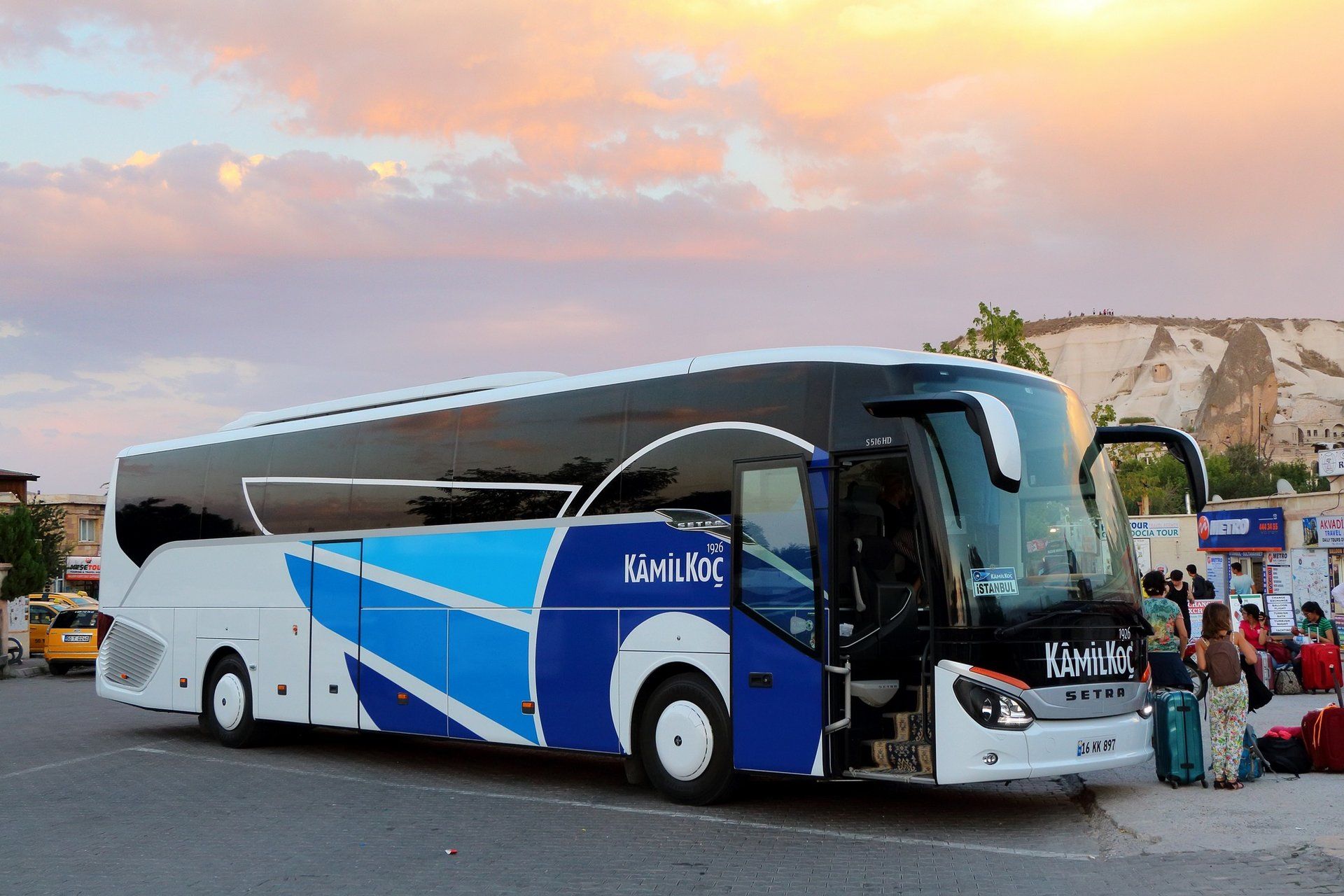Göreme'de gün batımı gökyüzü altında yolcu bekleyen mavi-beyaz şehirlerarası otobüs (Setra S 516 HD); ön kapı açık, sağda bavullu yolcular
