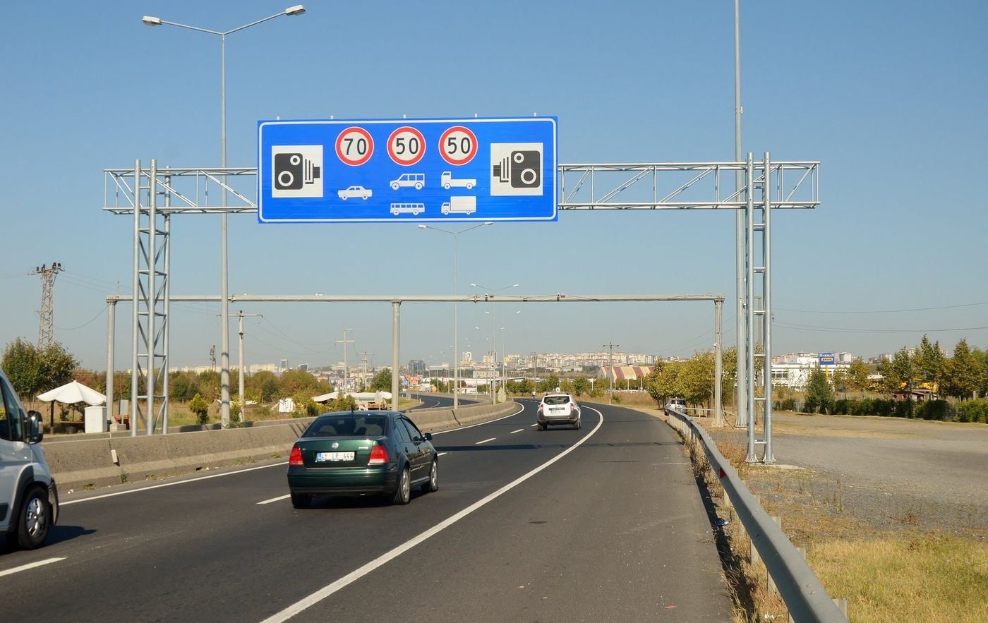 Türkiye'de D100 karayolu üzerinde araç sınıfı simgeleri ve hız sayıları taşıyan mavi portal tabela; altında seyreden otomobiller