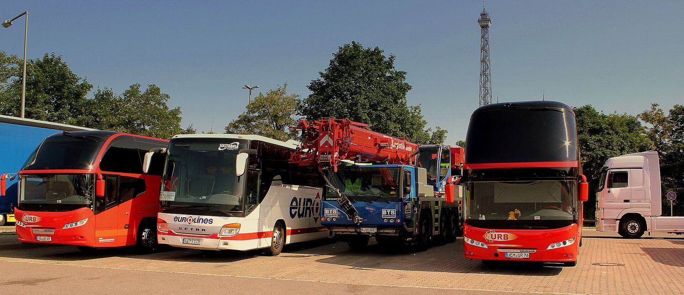 A red single-deck coach, a white coach, a blue truck-mounted mobile crane and a red double-deck coach parked side by side on a paved lot, with a white tractor unit at the right edge, the end of a blue trailer at the left edge, trees behind and a lattice radio tower against a clear blue sky