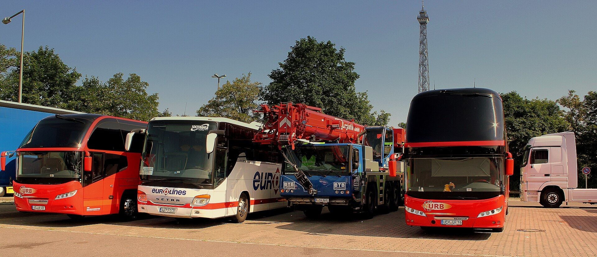 A red single-deck coach, a white coach, a blue truck-mounted mobile crane and a red double-deck coach parked side by side on a paved lot, with a white tractor unit at the right edge, the end of a blue trailer at the left edge, trees behind and a lattice radio tower against a clear blue sky