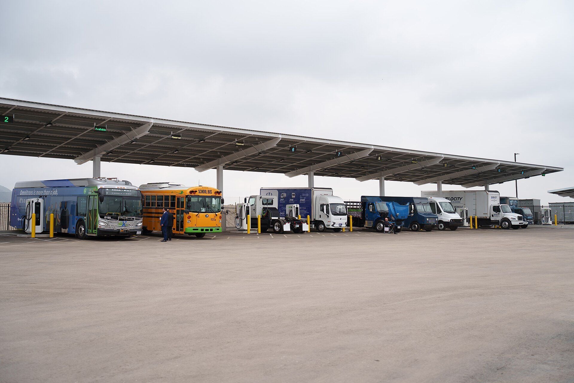 Electric city bus, school bus and several electric trucks parked side by side at charging bays under a long solar canopy