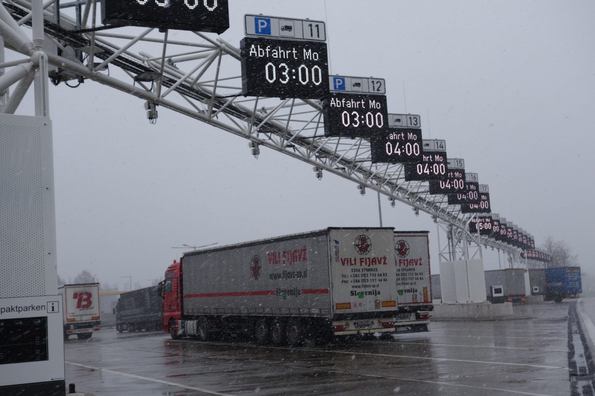 Trucks parked in rows at the Jura West rest area on the A3 near Velburg, Germany, under gantry displays showing departure times such as Abfahrt Mo 03:00 and Abfahrt Mo 04:00; a red tractor with a Slovenian curtainsider trailer in front, in falling snow