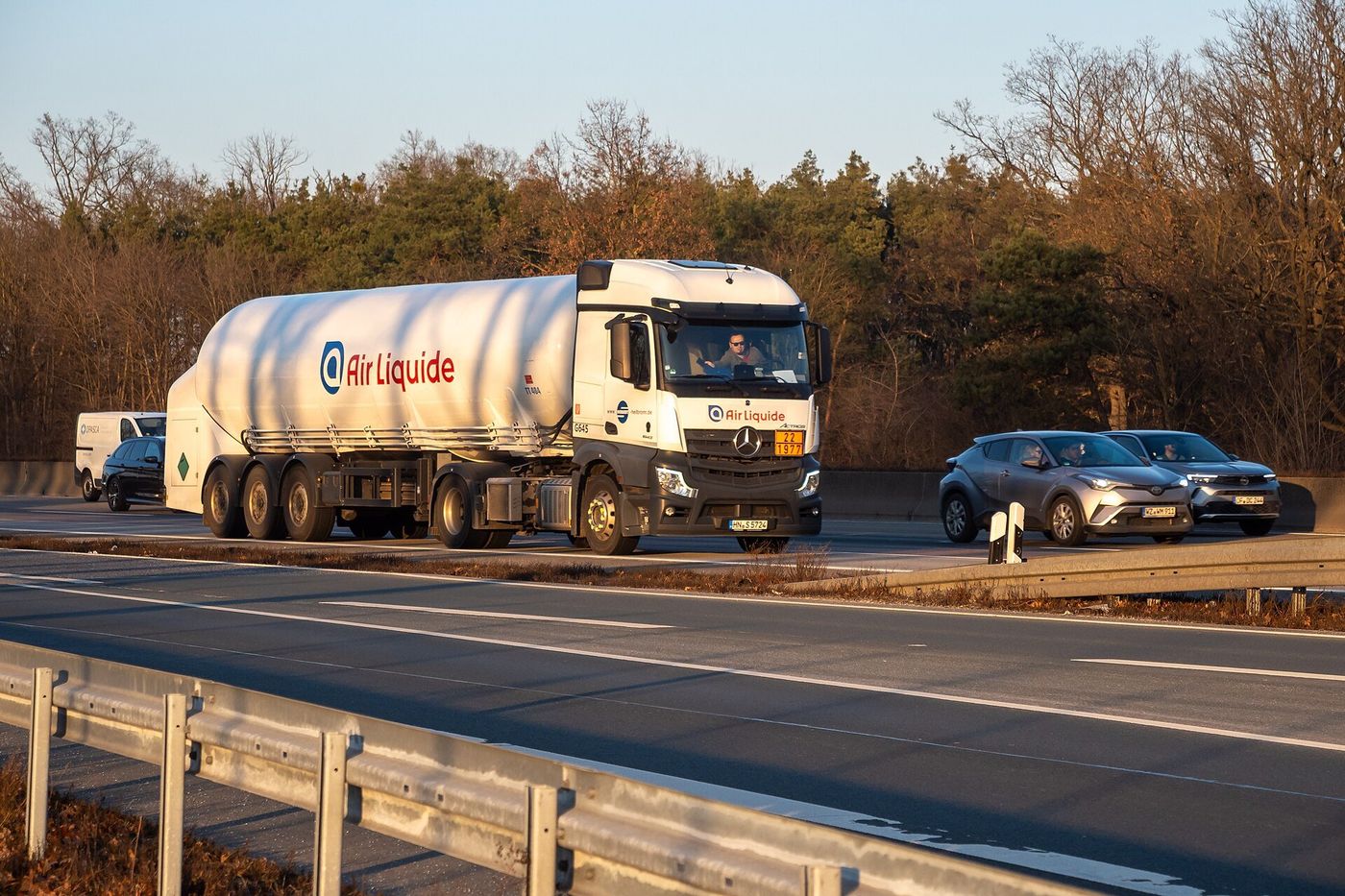 White Mercedes-Benz Actros tractor with an Air Liquide tank semi-trailer driving among cars on the A5 motorway near Frankfurt Airport, bare trees behind the crash barrier