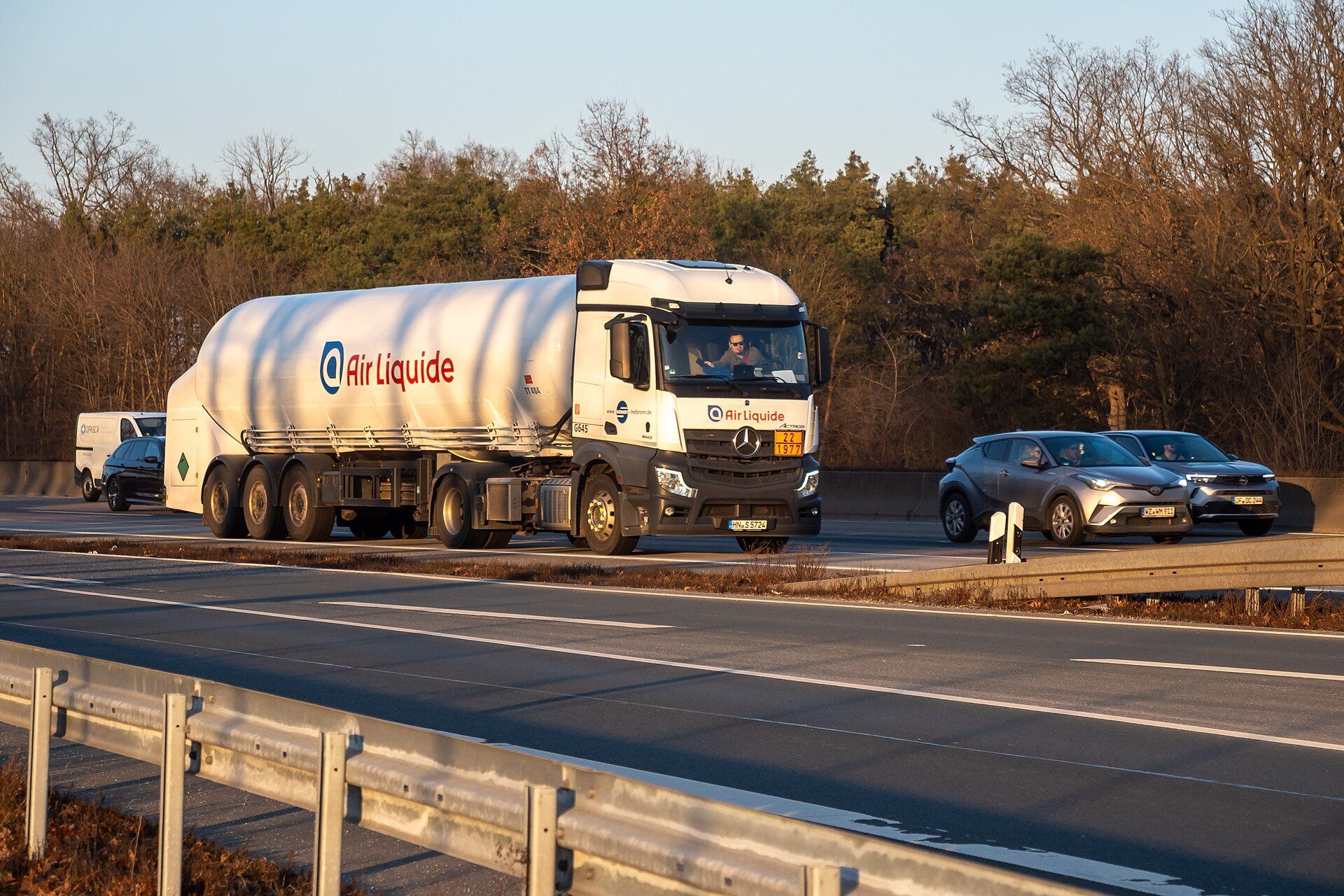 White Mercedes-Benz Actros tractor with an Air Liquide tank semi-trailer driving among cars on the A5 motorway near Frankfurt Airport, bare trees behind the crash barrier
