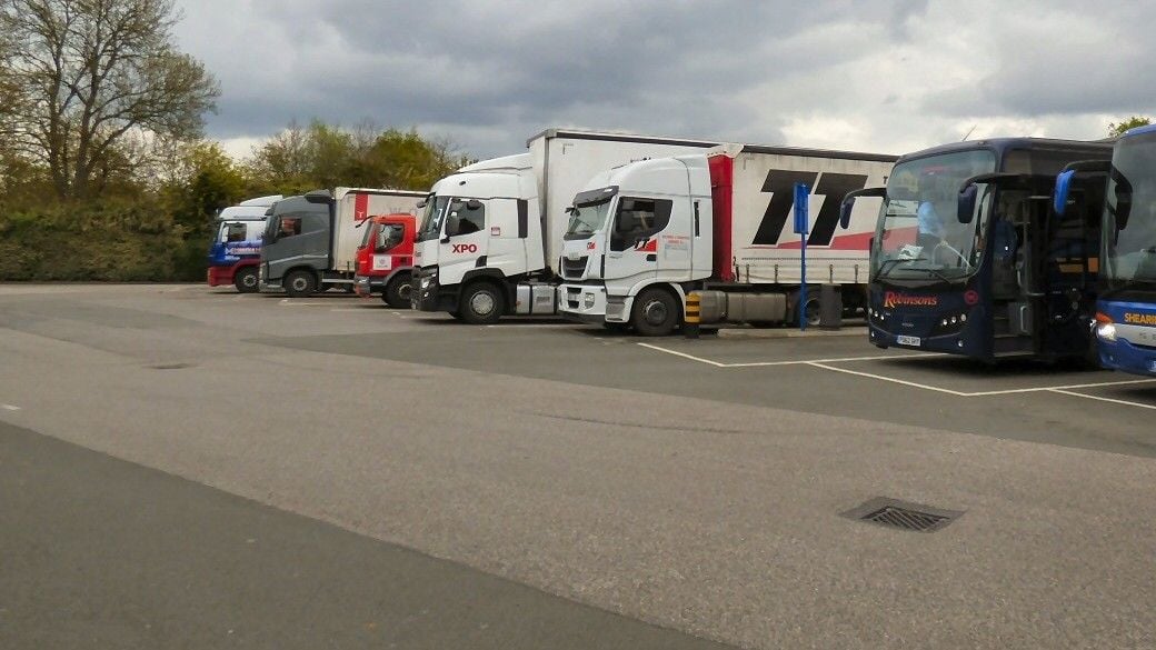 A row of lorries, most of them tractor units with box or curtain-sided semi-trailers, parked beside a coach and the front of a second coach in a lorry and coach park under a cloudy sky