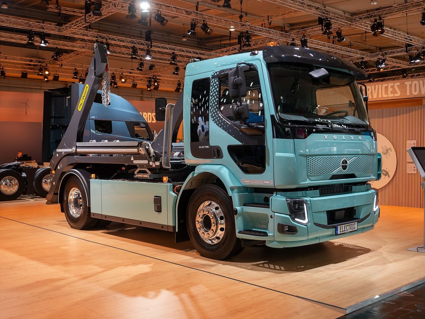 Turquoise Volvo FE Electric two-axle rigid with the current front end, V-shaped LED headlights and a Palfinger skip loader, on a trade-fair stand at IAA Transportation 2024 in Hanover