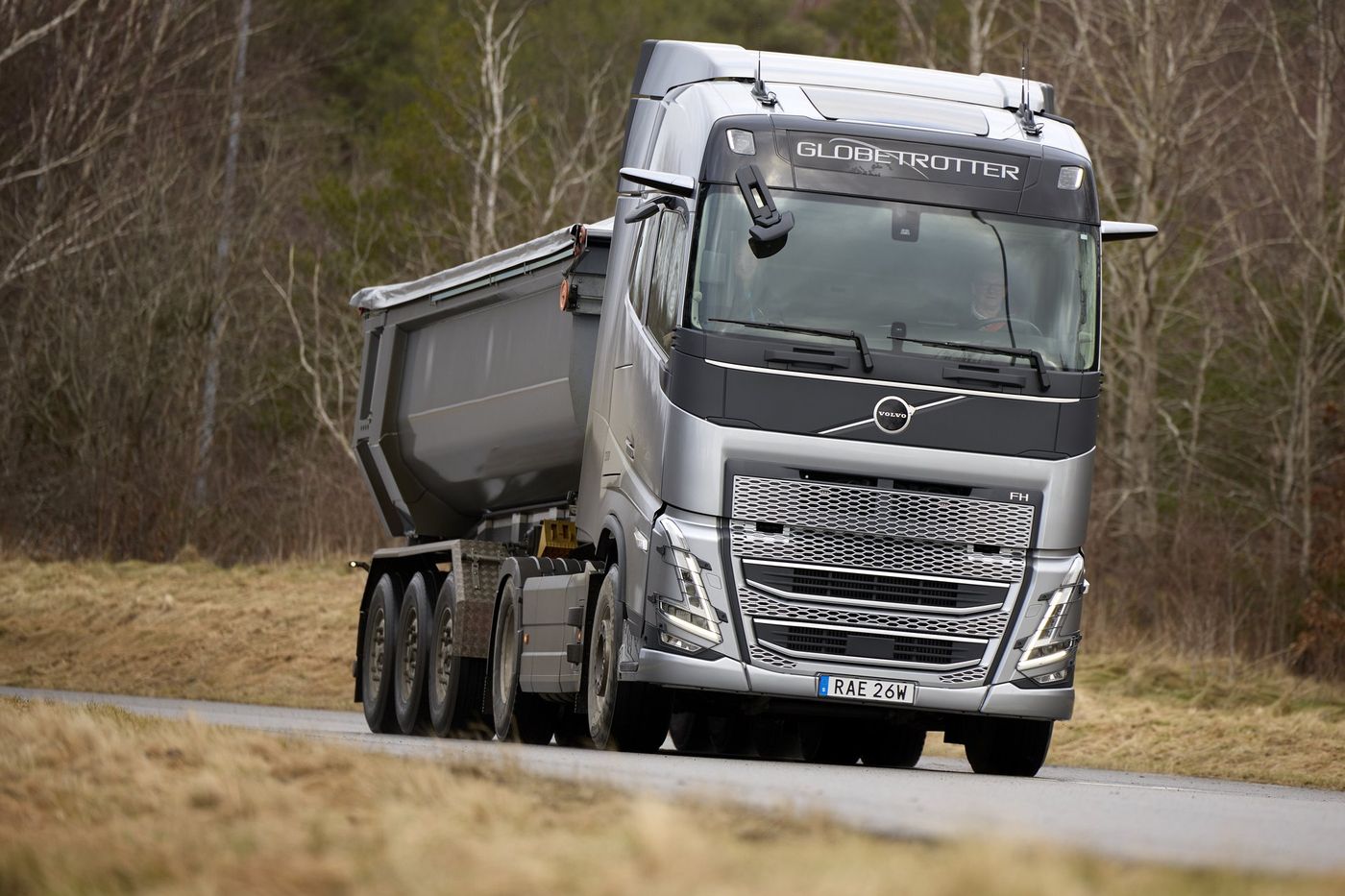 Silver 2020-generation Volvo FH two-axle tractor unit with a Globetrotter cab, pulling a three-axle tipper semi-trailer along a road through bare winter woodland
