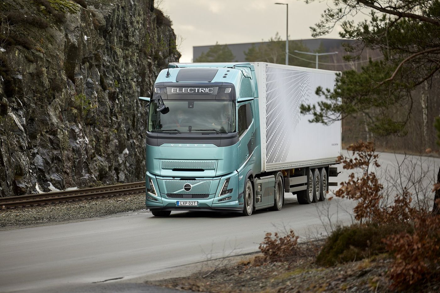 Light blue-green Volvo FH Aero Electric tractor unit with the word ELECTRIC above the windscreen, pulling a white box semitrailer along a road beside a rock face