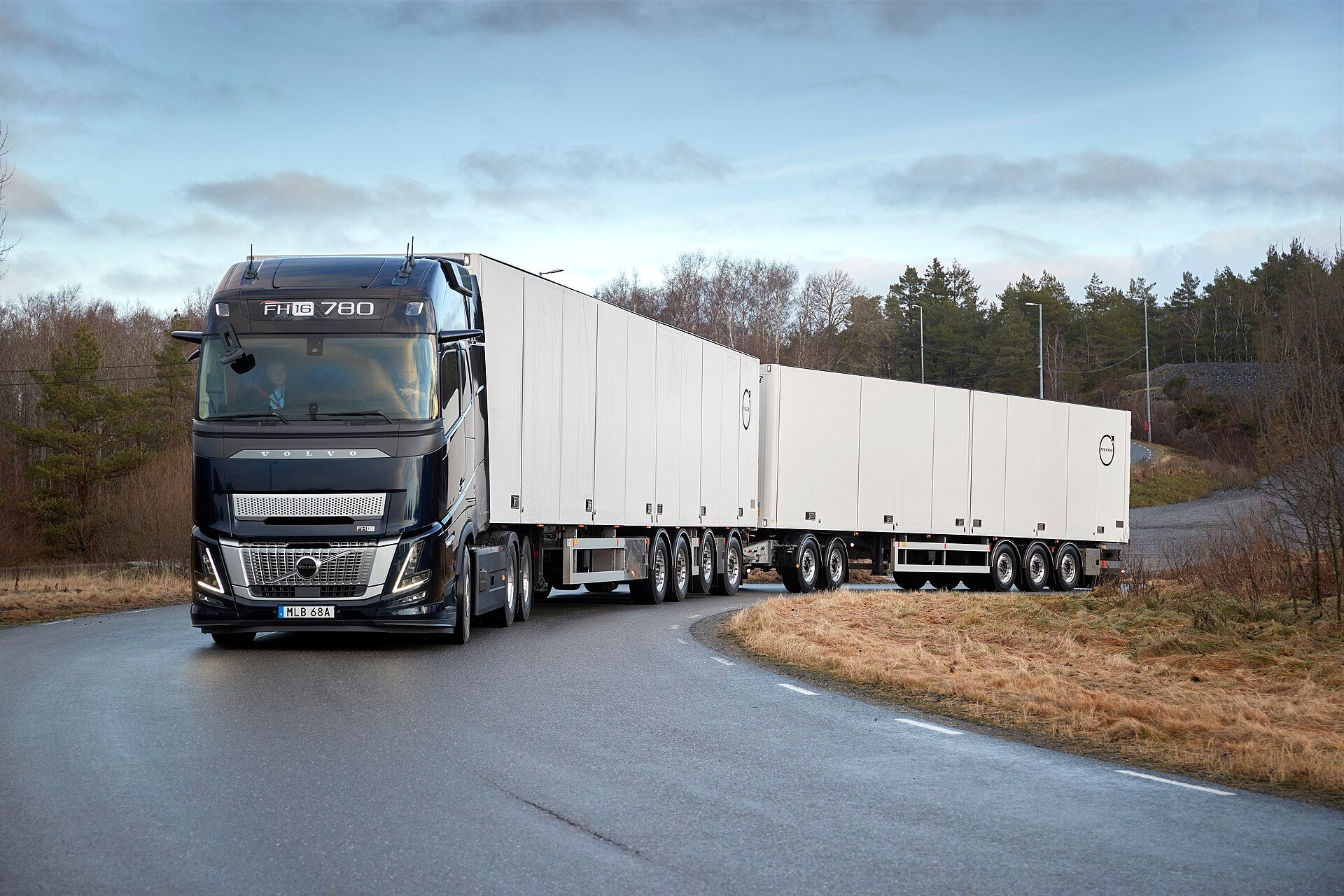 Black Volvo FH16 three-axle tractor unit with an extended front carrying VOLVO lettering and an FH16 780 sign above the windscreen, pulling two white box semi-trailers in a double combination round a bend on a wet road