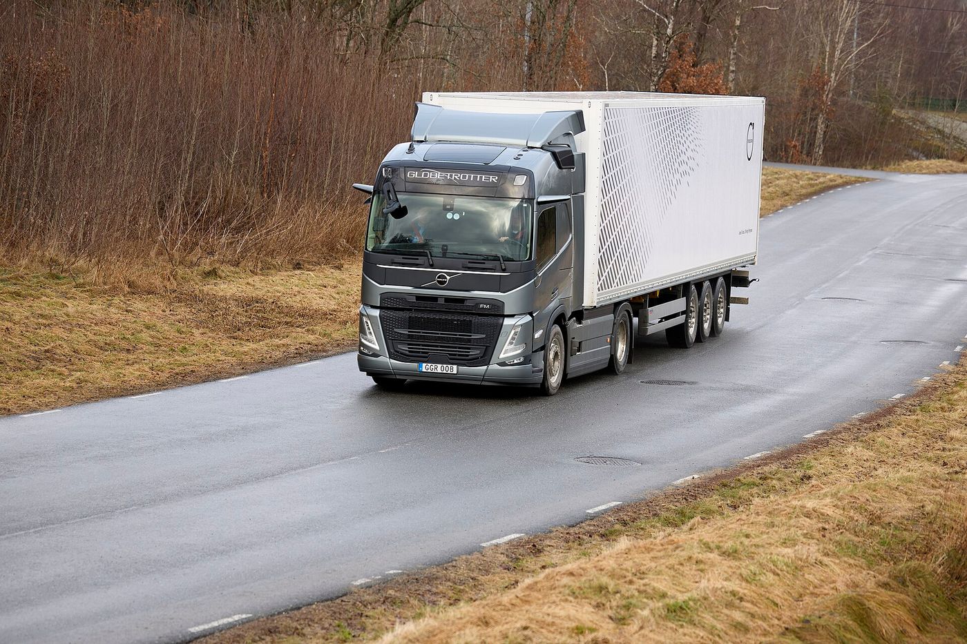 Dark grey 2020-generation Volvo FM two-axle tractor unit with a Globetrotter cab, pulling a white three-axle box semi-trailer along a wet road through bare winter woodland