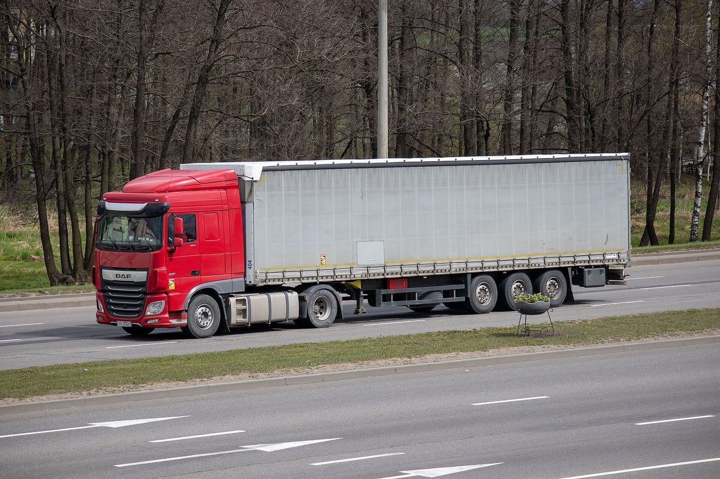 Red two-axle DAF tractor unit pulling a plain grey curtain-sided semi-trailer with three axles along a multi-lane road, seen from the front left, with bare trees behind