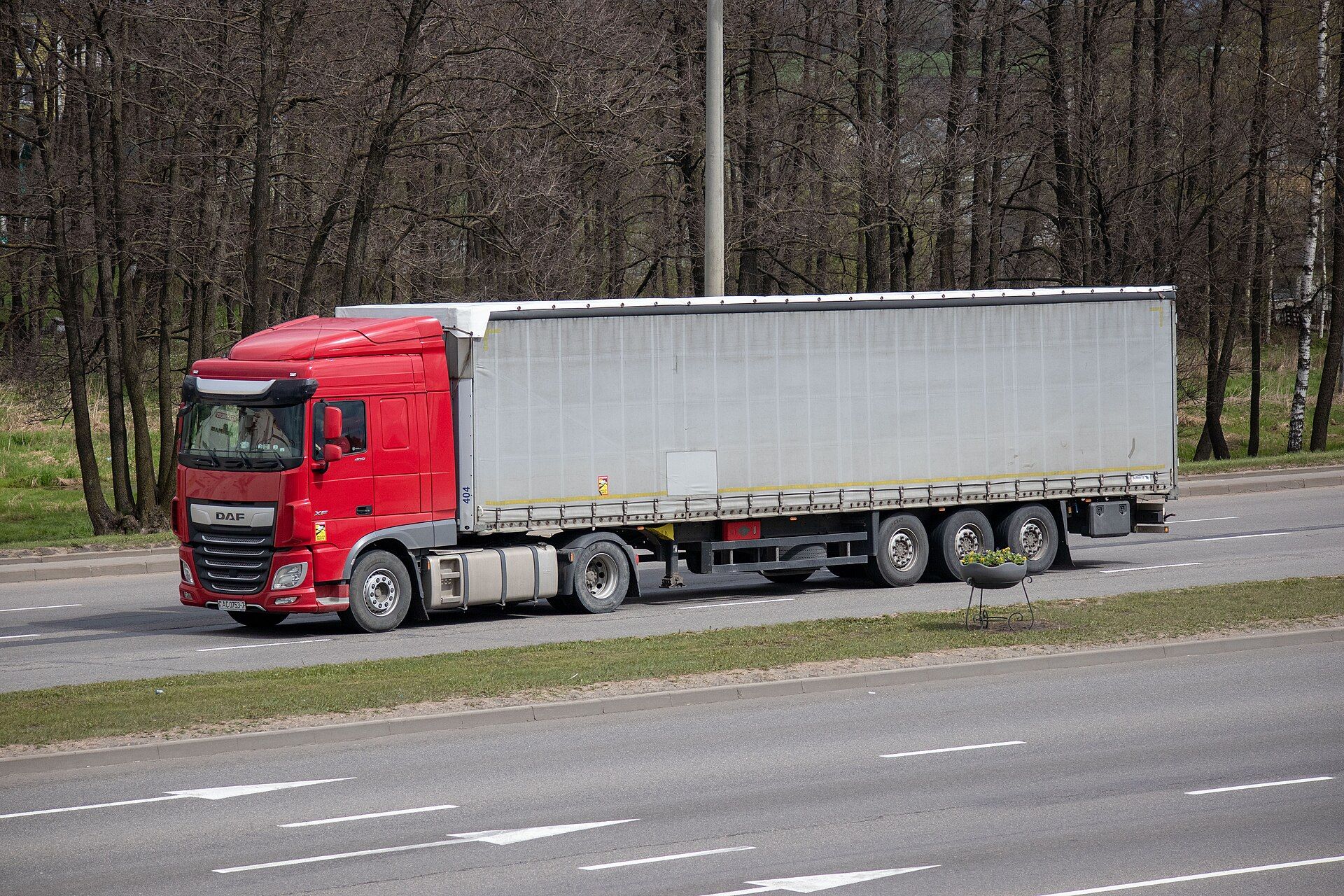 Red two-axle DAF tractor unit pulling a plain grey curtain-sided semi-trailer with three axles along a multi-lane road, seen from the front left, with bare trees behind