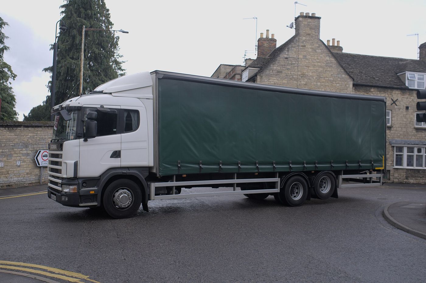 A white Scania rigid lorry with a plain green curtain-sided body on three axles turns at a junction in front of stone houses; at the left, a road sign shows a red-ringed lorry symbol marked 7.5T