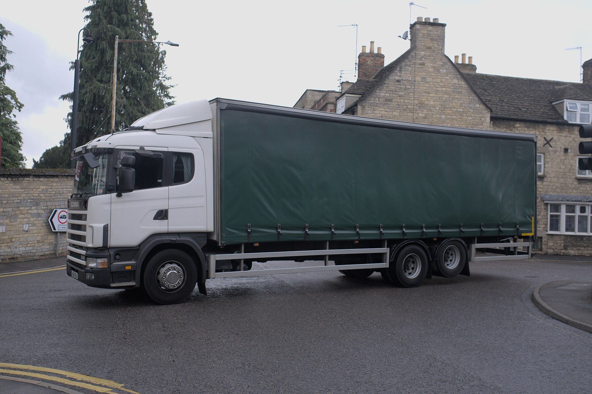A white Scania rigid lorry with a plain green curtain-sided body on three axles turns at a junction in front of stone houses; at the left, a road sign shows a red-ringed lorry symbol marked 7.5T