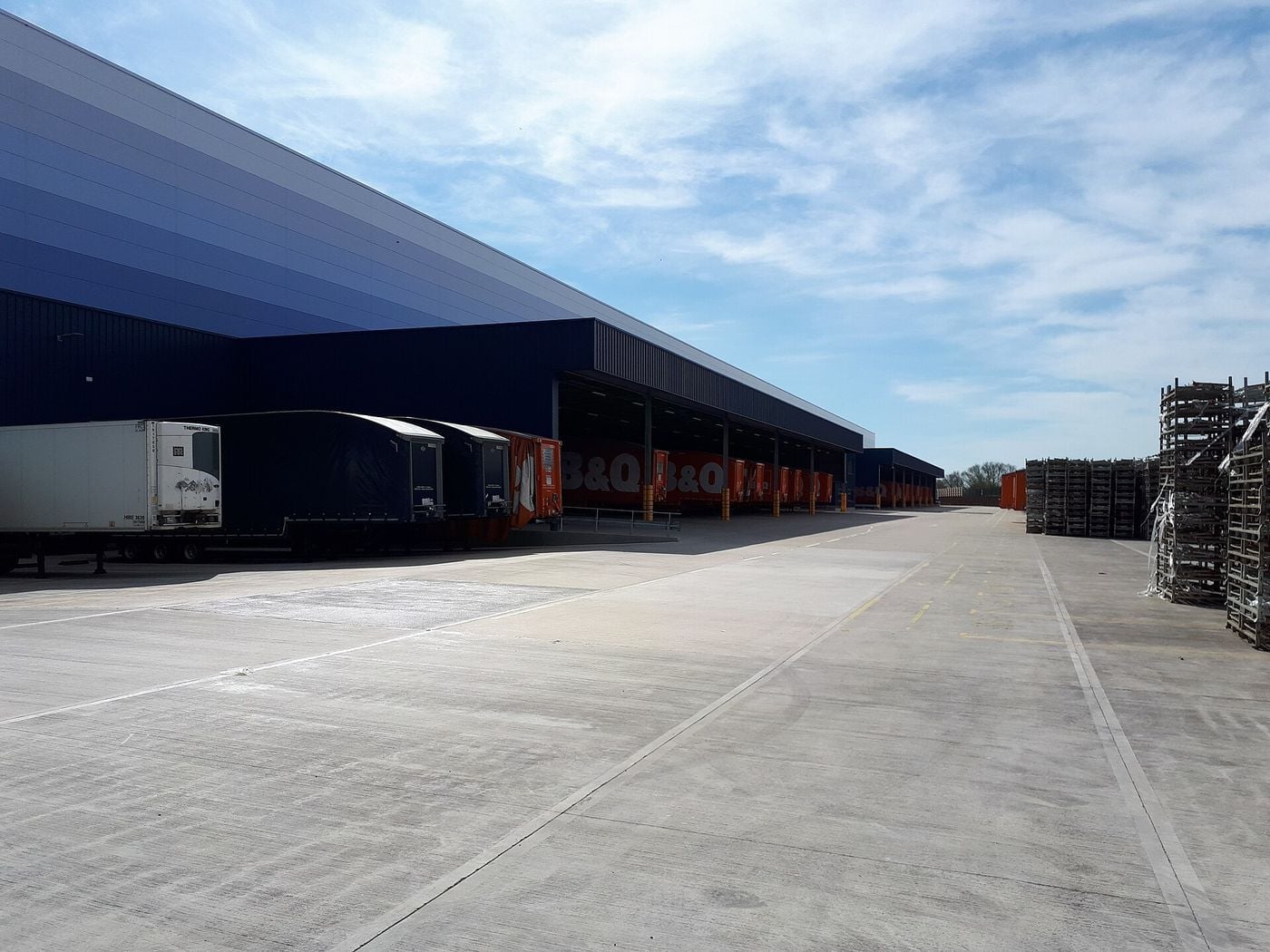Refrigerated, curtain-sided and box semi-trailers backed up to the loading doors of a large distribution centre in Swindon, England, with empty roll cages stacked on the concrete yard